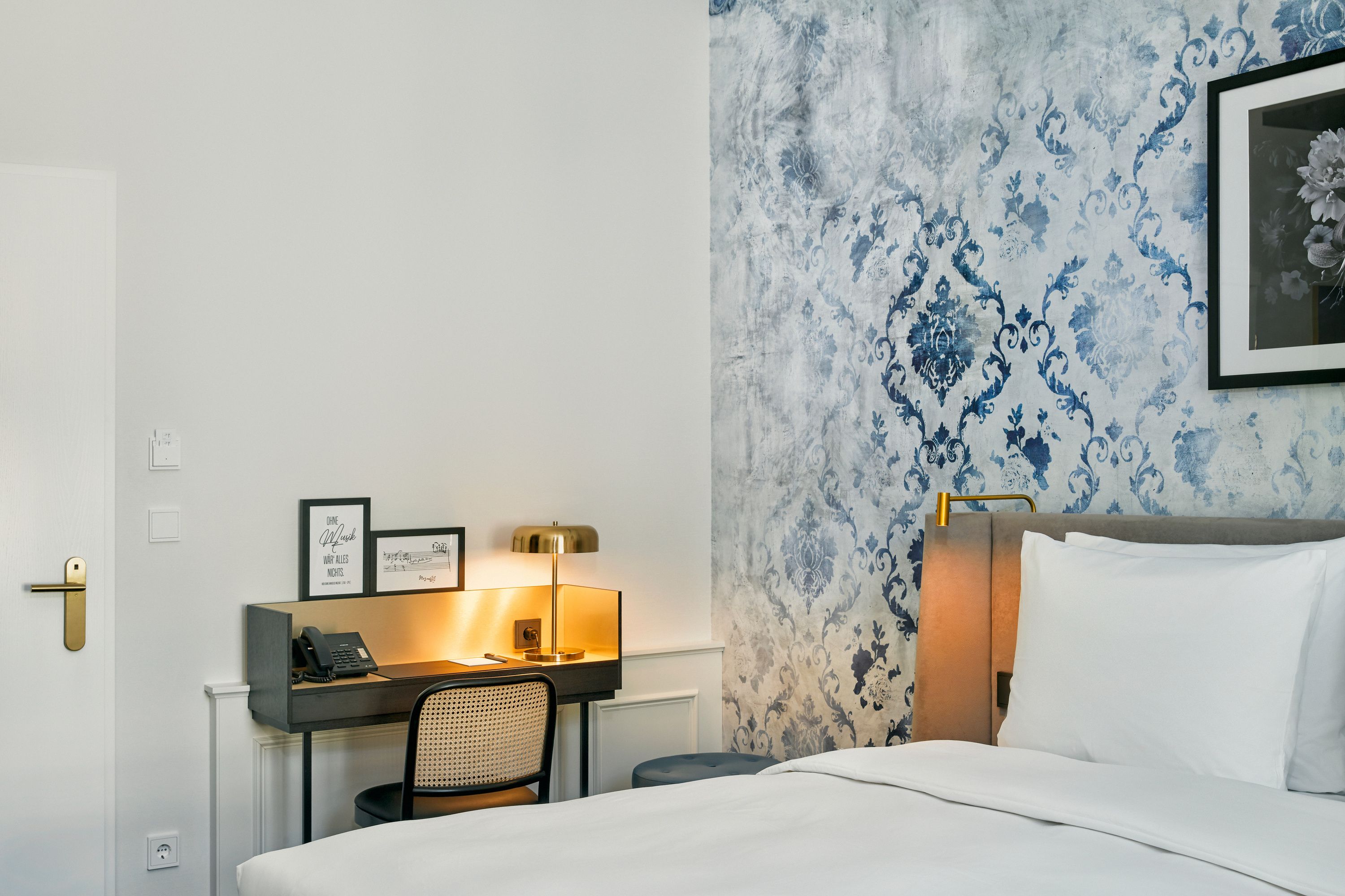 Helles Hotelzimmer mit gemütlichem Doppelbett, stilvoll gemusterter Wand, Schreibtisch mit Retro-Lampe und Telefon, ruhige Atmosphäre ideal für entspanntes Arbeiten und erholsamen Schlaf.