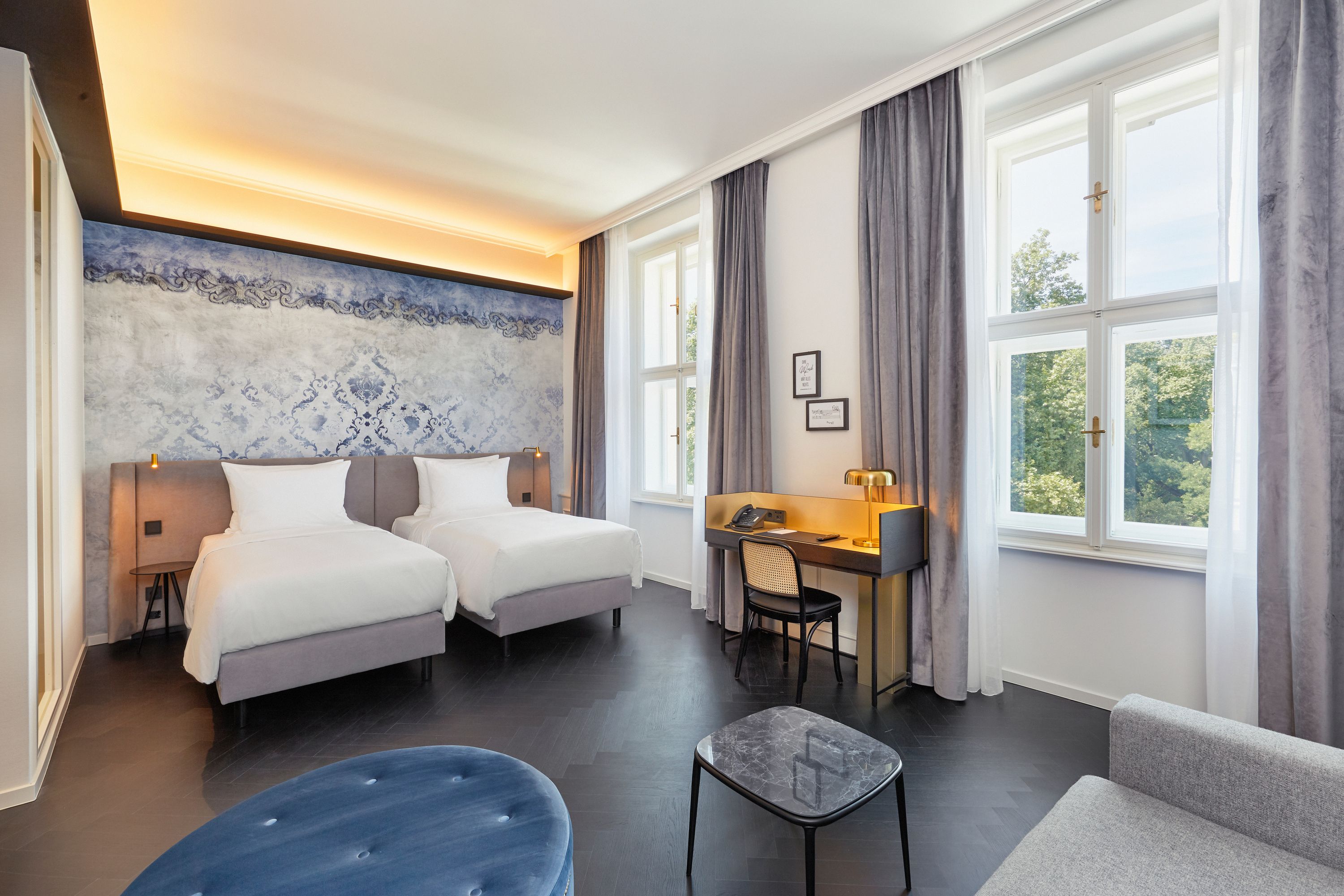 Modern eingerichtetes Hotelzimmer mit zwei Einzelbetten, großem Fenster mit Tageslicht, Schreibtisch mit Lampe und gemütlicher Sitzecke, das eine entspannte Atmosphäre für Geschäfts- und Freizeitreisende bietet.