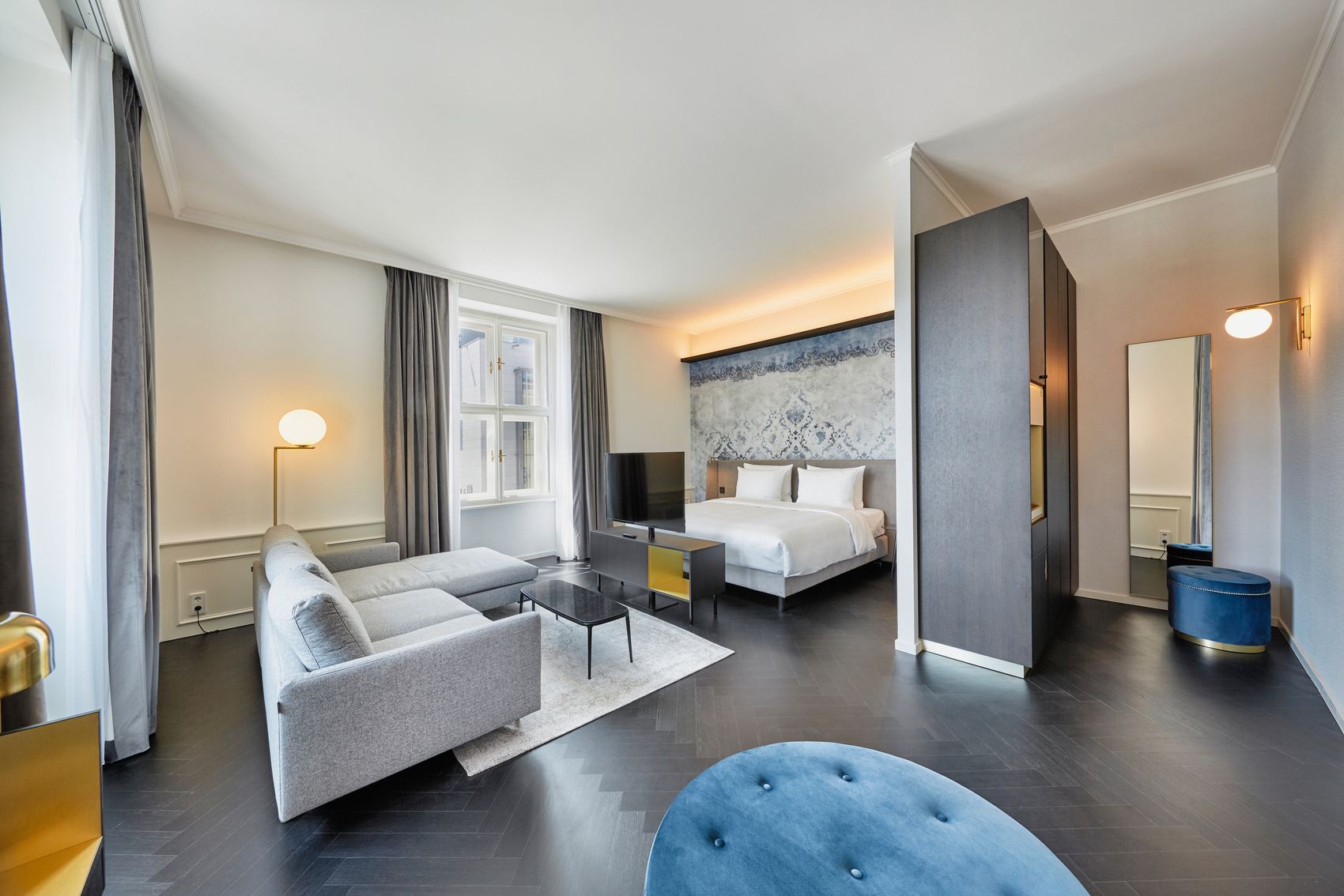 Helles, modernes Hotelzimmer mit großem Doppelbett, gemütlicher Sitzecke mit Sofa, Flachbildfernseher und großem Fenster für viel Tageslicht. Zeitgemäßes Design in neutralen Farben schafft ein einladendes Ambiente für entspannte Aufenthalte.