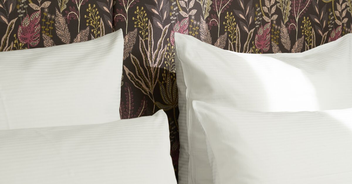 Weiche, weiße Kissen vor einer Wand mit floralem, elegantem Muster, die Behaglichkeit und stilvolles Ambiente im Zimmer vermittelt.