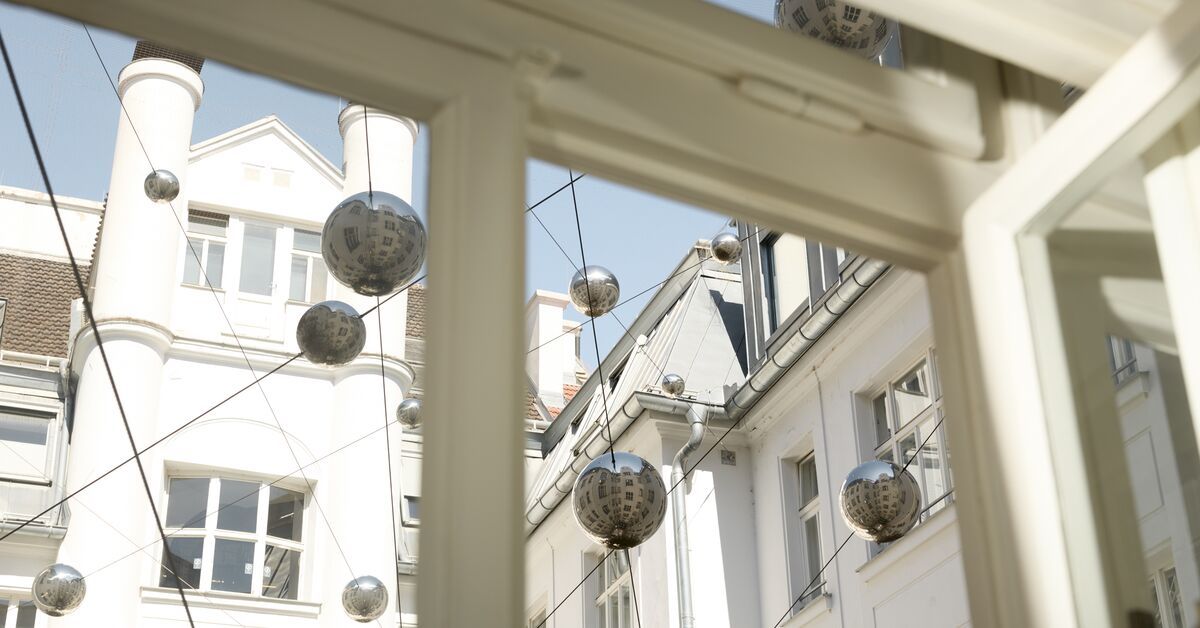 Blick aus einem Fenster auf eine ruhige Innenhofszene mit historischen weiß gestrichenen Gebäuden und dekorativen, glänzenden Kugeln, die an Drähten zwischen den Häusern hängen. Das Licht vermittelt eine helle, freundliche Atmosphäre.