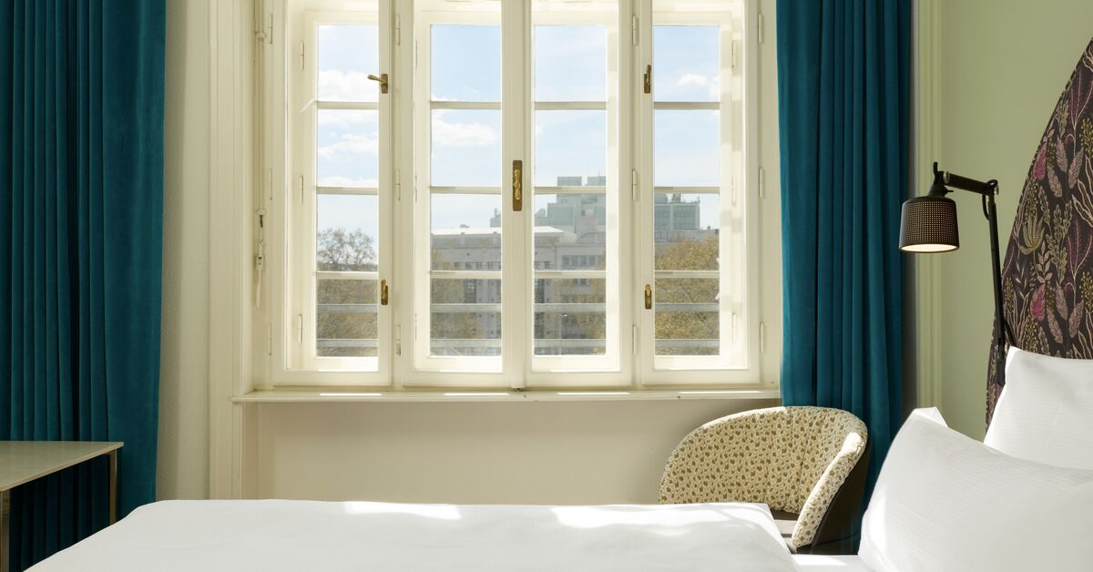 Helles Hotelzimmer mit großem Fenster, das Tageslicht hereinlässt. Gemütlicher Sessel neben dem Bett mit weißen Bettwäsche und dunkelblauen Vorhängen für eine entspannte Atmosphäre.