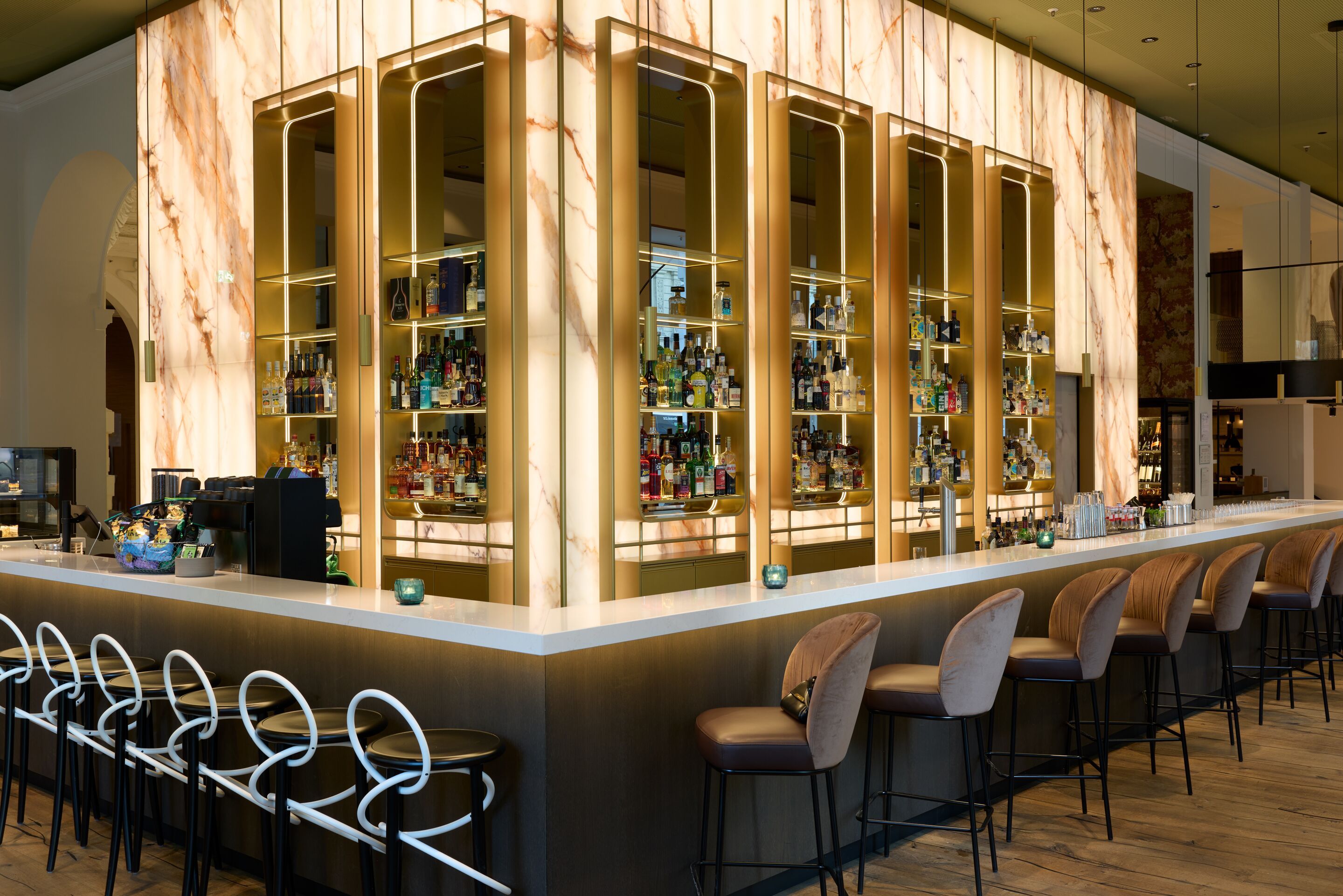 Elegante Hotelbar mit beleuchtetem Marmorelement, modernen Barhockern und einer Vielzahl an Getränken, die in Regalen hinter dem Tresen präsentiert werden. Einladende Atmosphäre für entspannte Abende.