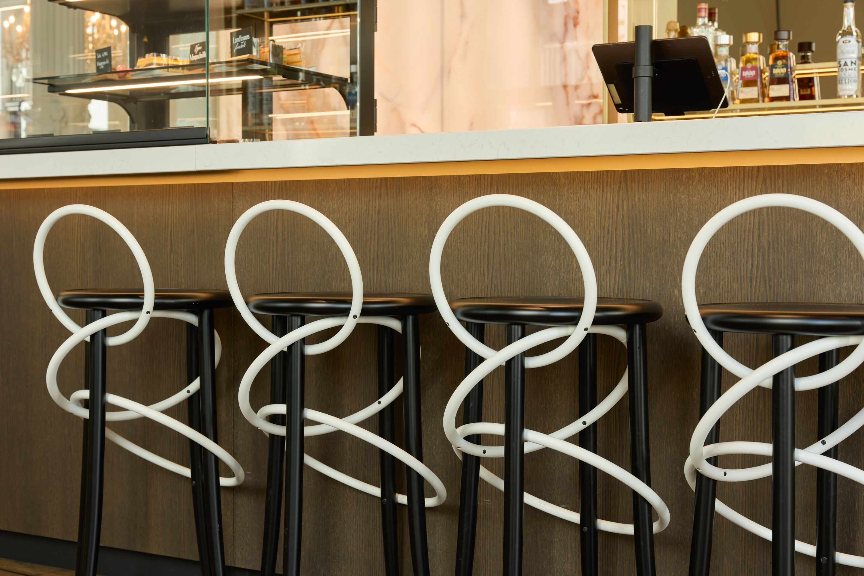 Moderne Barhocker mit weißen, geschwungenen Rückenlehnen vor einer stilvollen Bar, die Getränke und Gebäck präsentiert. Ambiente wirkt einladend und zeitgemäß, ideal für entspannte Stunden in der Hotelbar.