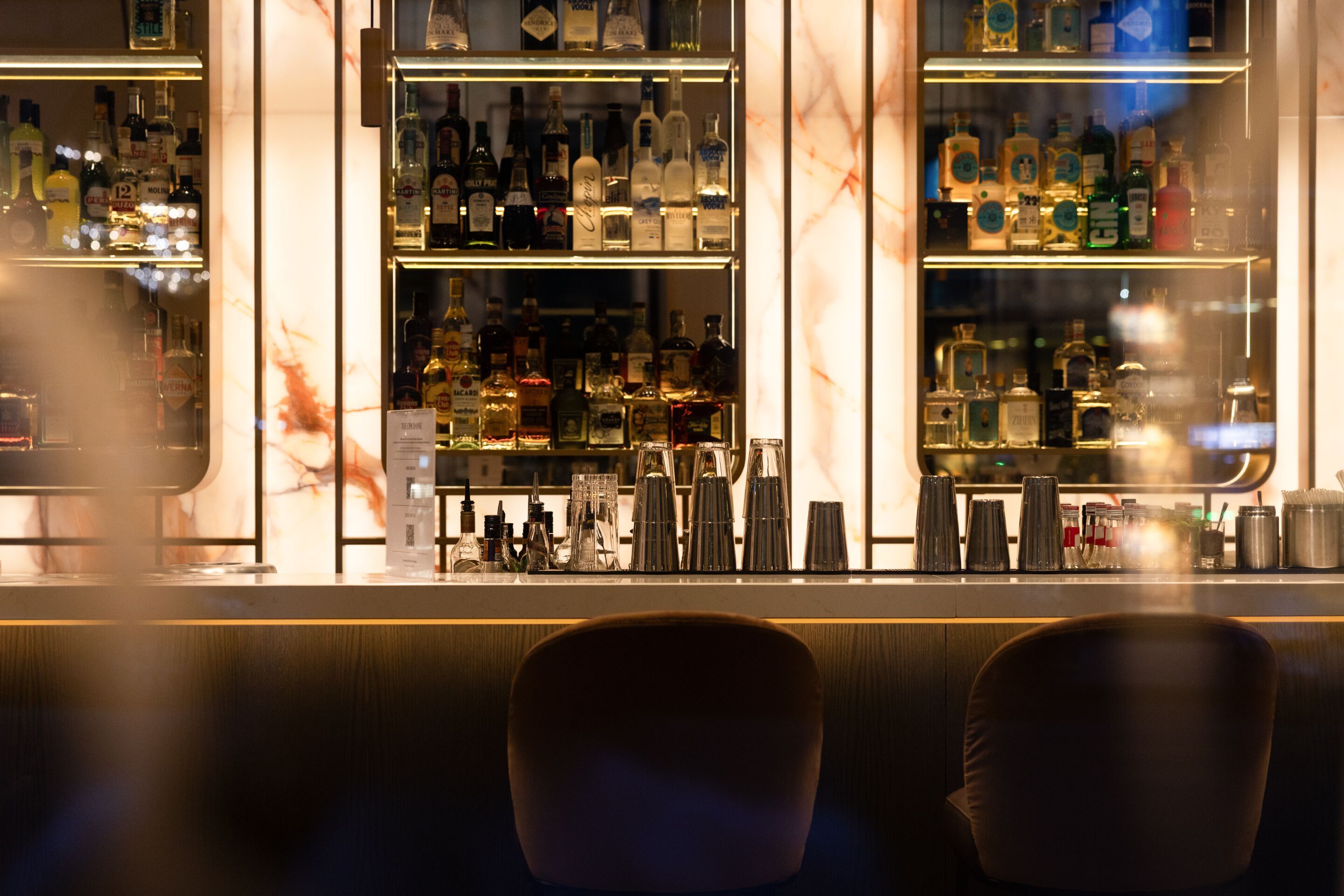 Elegante Hotelbar mit beleuchteten Regalen voller Spirituosen, modernem Tresen und zwei bequemen Barhockern in gedämpfter, einladender Atmosphäre.