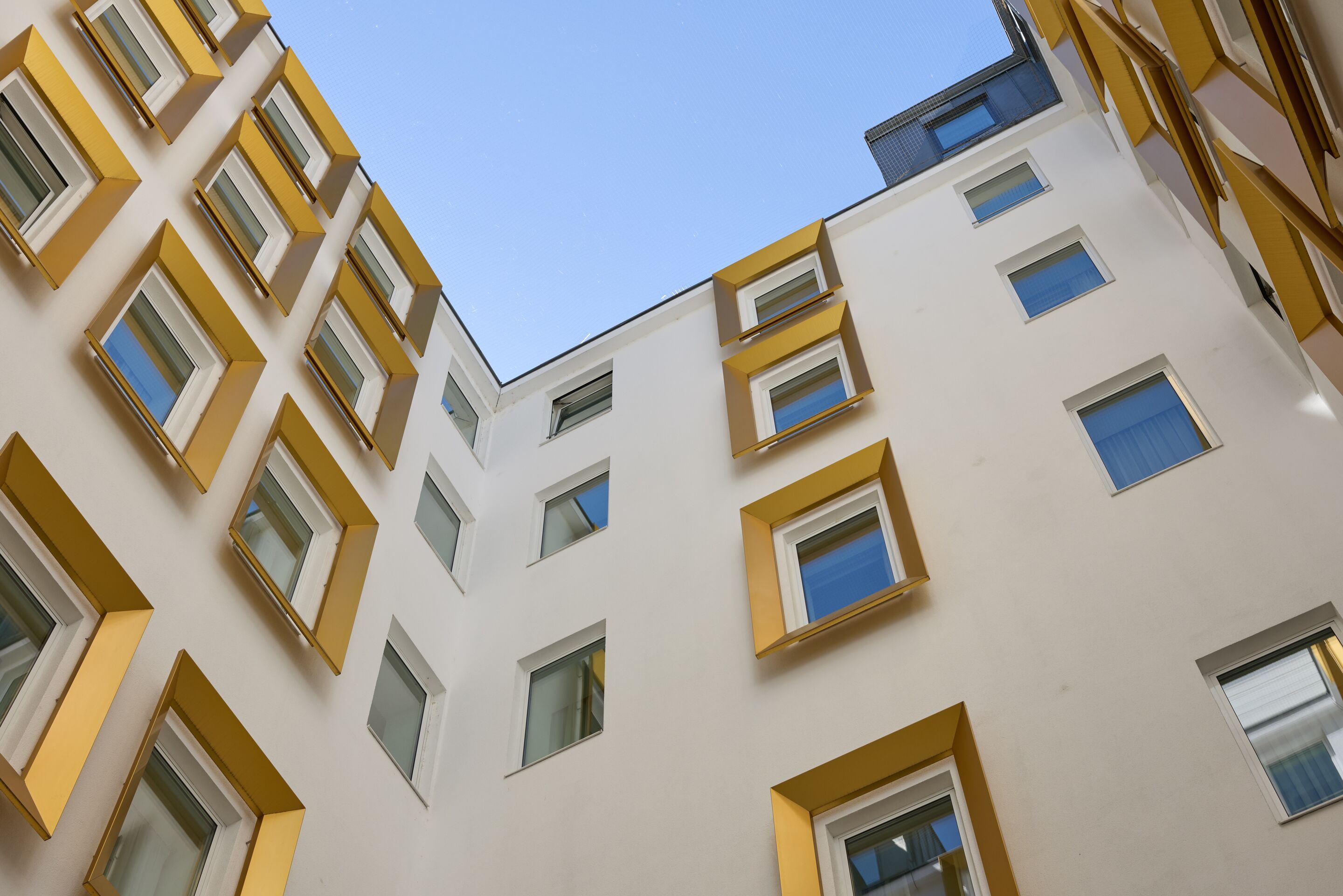 Blick in einen Innenhof eines modernen Hotels mit weißen Fassaden und Fensterrahmen in Gelb, die den klaren blauen Himmel einrahmen.
