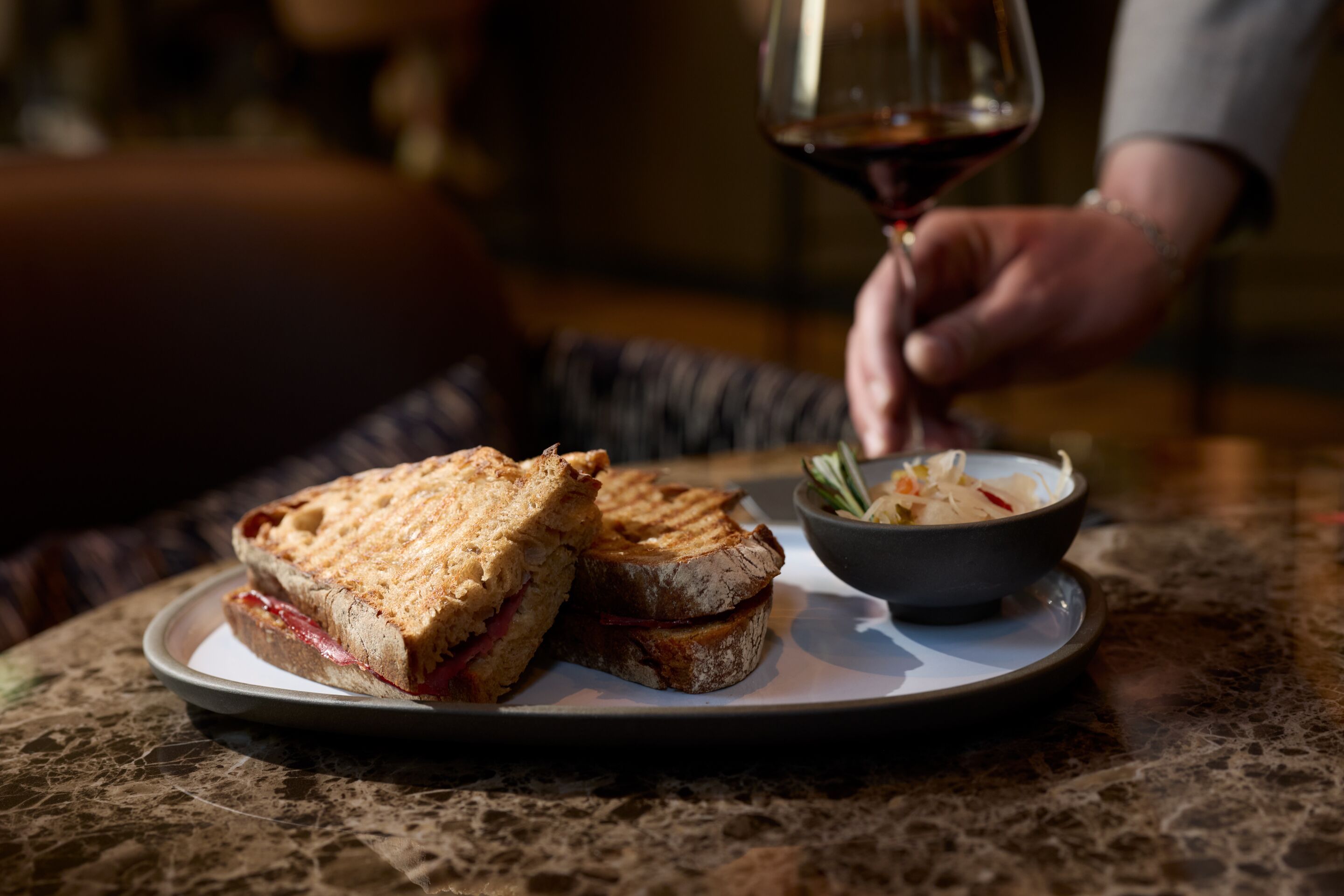 Auf einem Tisch steht ein Snack-Teller mit gegrilltem Sandwich und einer kleinen Schale Dip, daneben hält eine Hand ein Glas Rotwein. Die Szene strahlt gemütliche Genussmomente in entspannter Atmosphäre aus.