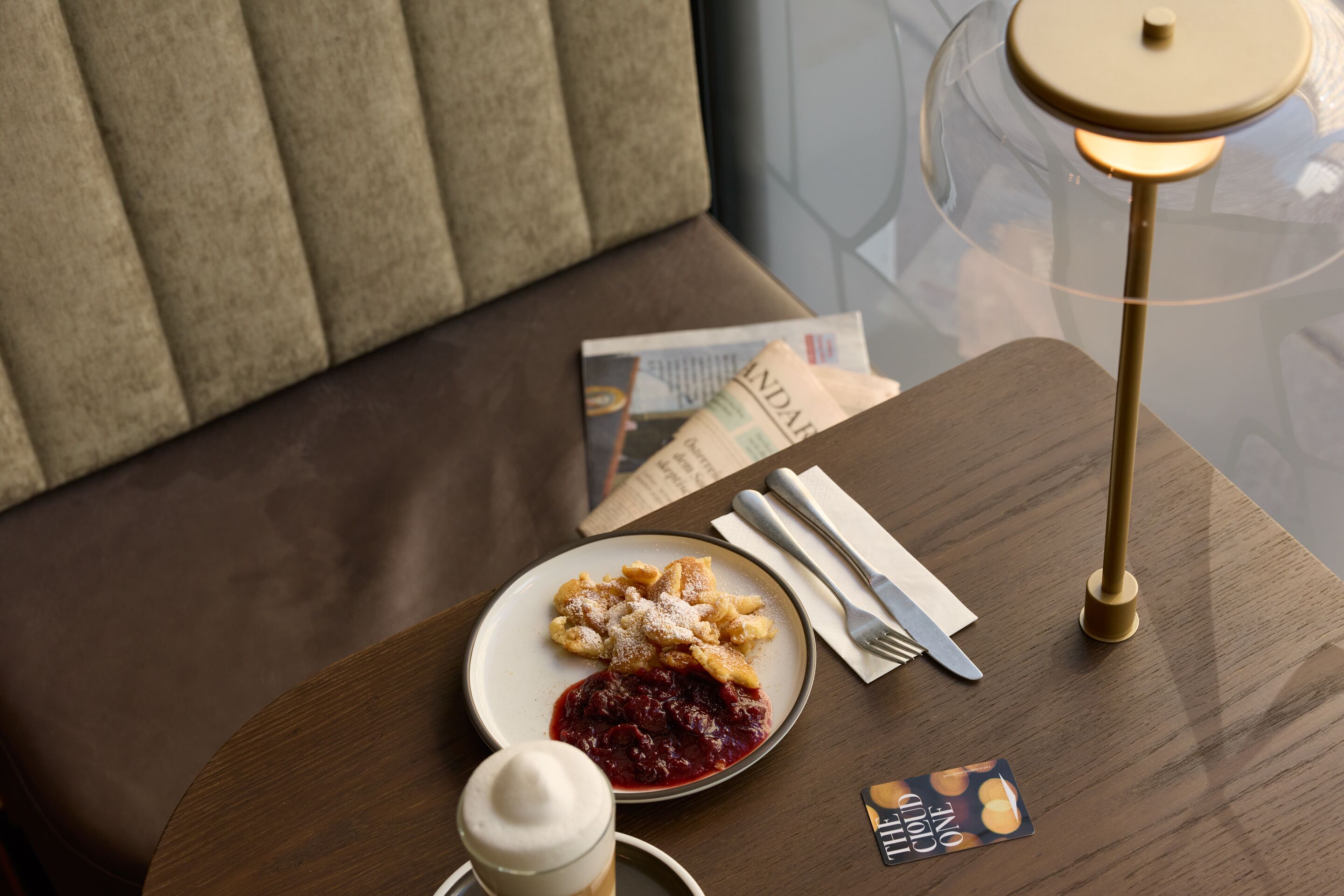 Gemütliche Sitzecke mit einem Tisch, darauf ein Teller mit Kaiserschmarrn und Preiselbeeren, ein Kaffee mit Milchschaum sowie Besteck und eine stilvolle Lampe, ideal für eine entspannte Pause oder ein Frühstück im Hotel.