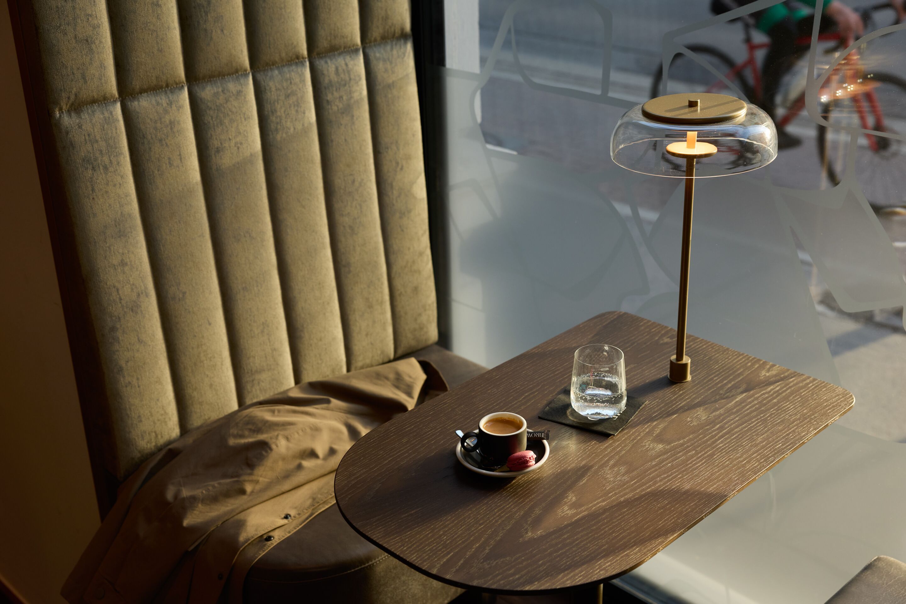 Gemütliche Sitzecke mit samtbezogenem Sessel, Holztisch, Espresso in Tasse, Glas Wasser und stilvoller Tischlampe vor großem Fenster mit Blick ins Freie.