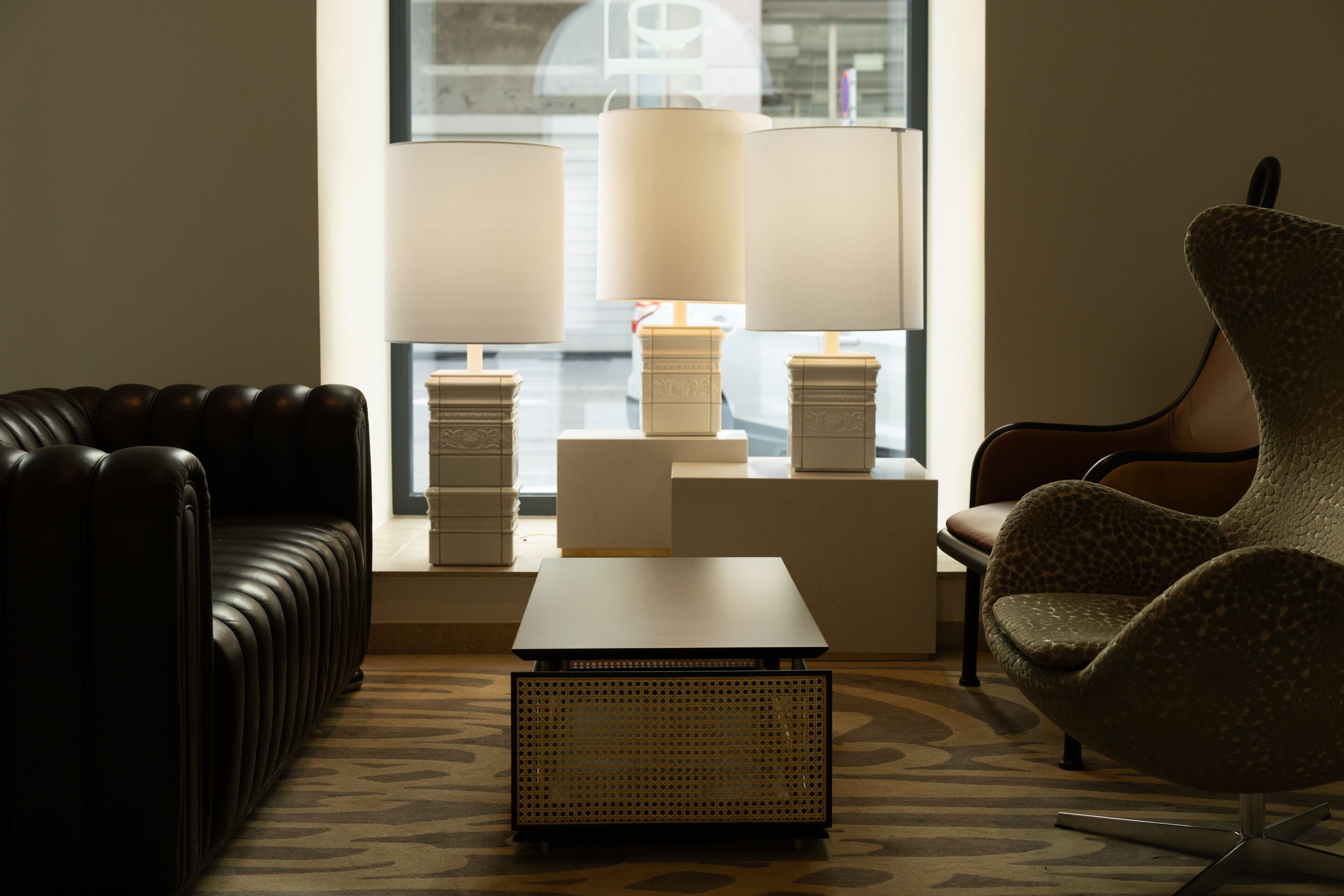 Gemütliche Sitzecke mit zwei unterschiedlich gestalteten Sesseln, einem schwarzen Ledersofa, einem kleinen Tisch und drei modernen Lampen vor einem großen Fenster, das sanftes Tageslicht hereinlässt. Eine einladende Atmosphäre für entspannte Gespräche oder ruhige Momente.