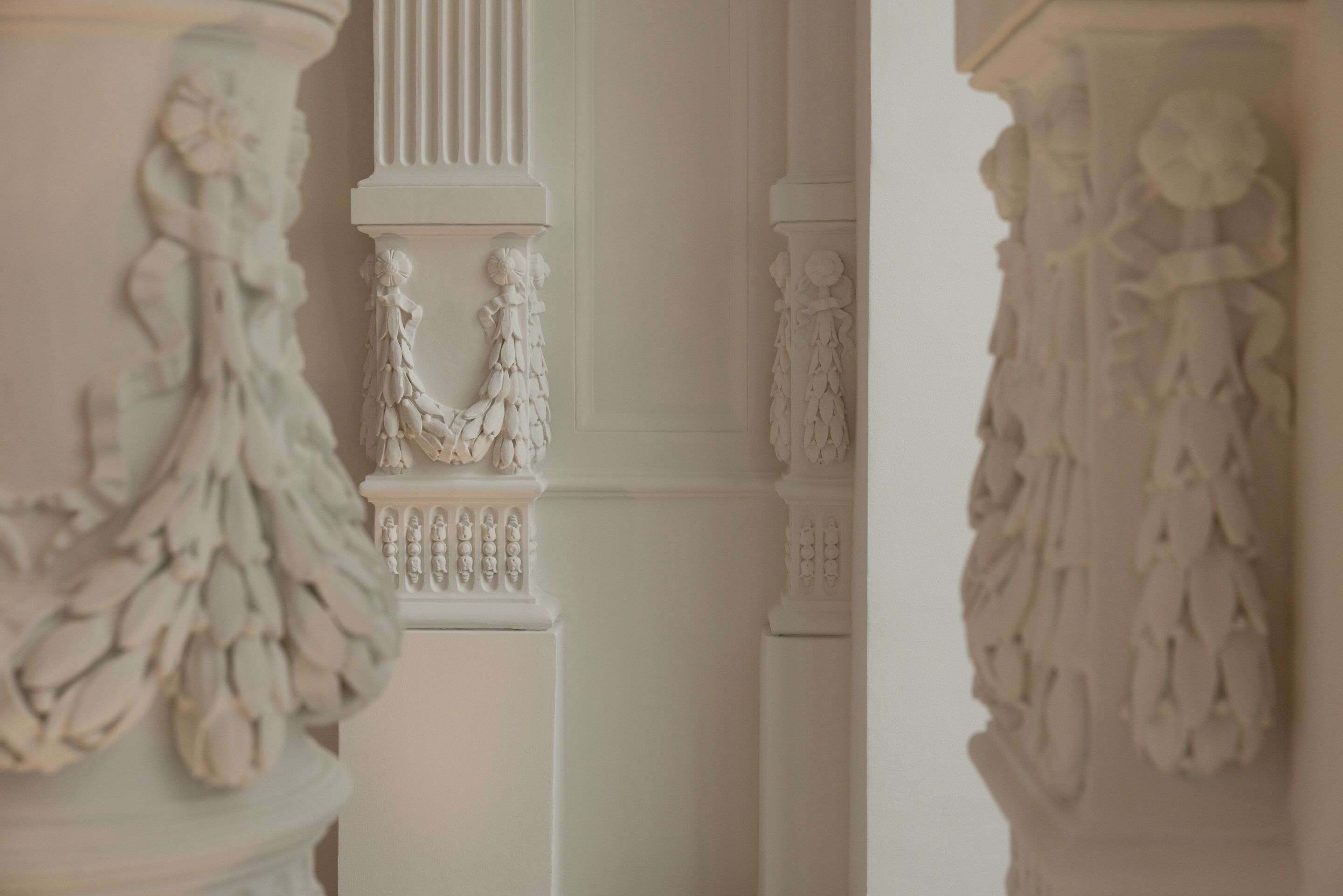Detailaufnahme von eleganten weißen Säulen mit kunstvollen, floralen Verzierungen in einem klassischen Innenraum, der eine ruhige und stilvolle Atmosphäre vermittelt.