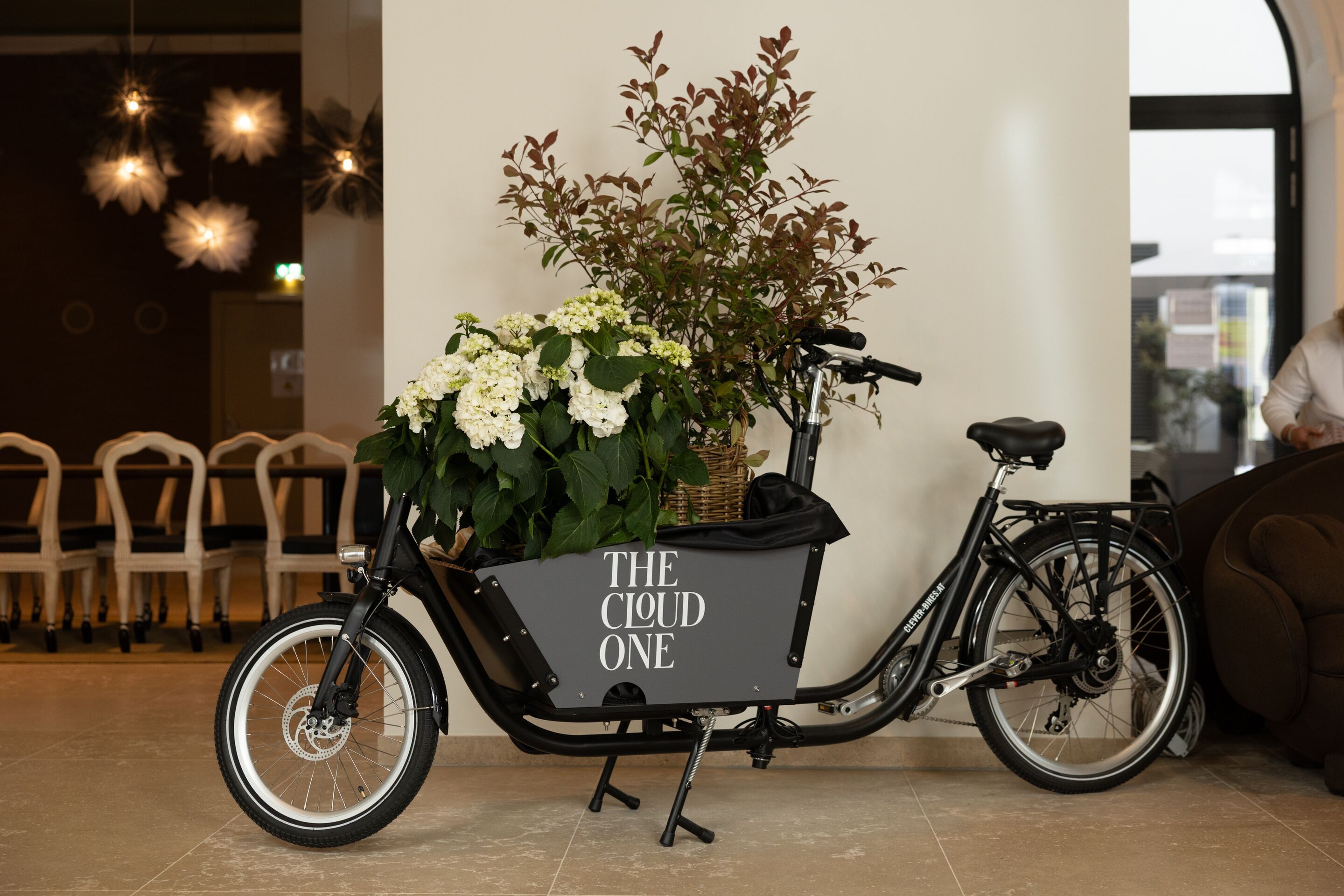Lastenfahrrad mit weißen Blumen und grünem Blattwerk, steht in einem hellen Innenraum neben einer Sitzgruppe. Auf der Ladefläche steht „THE CLOUD ONE“.