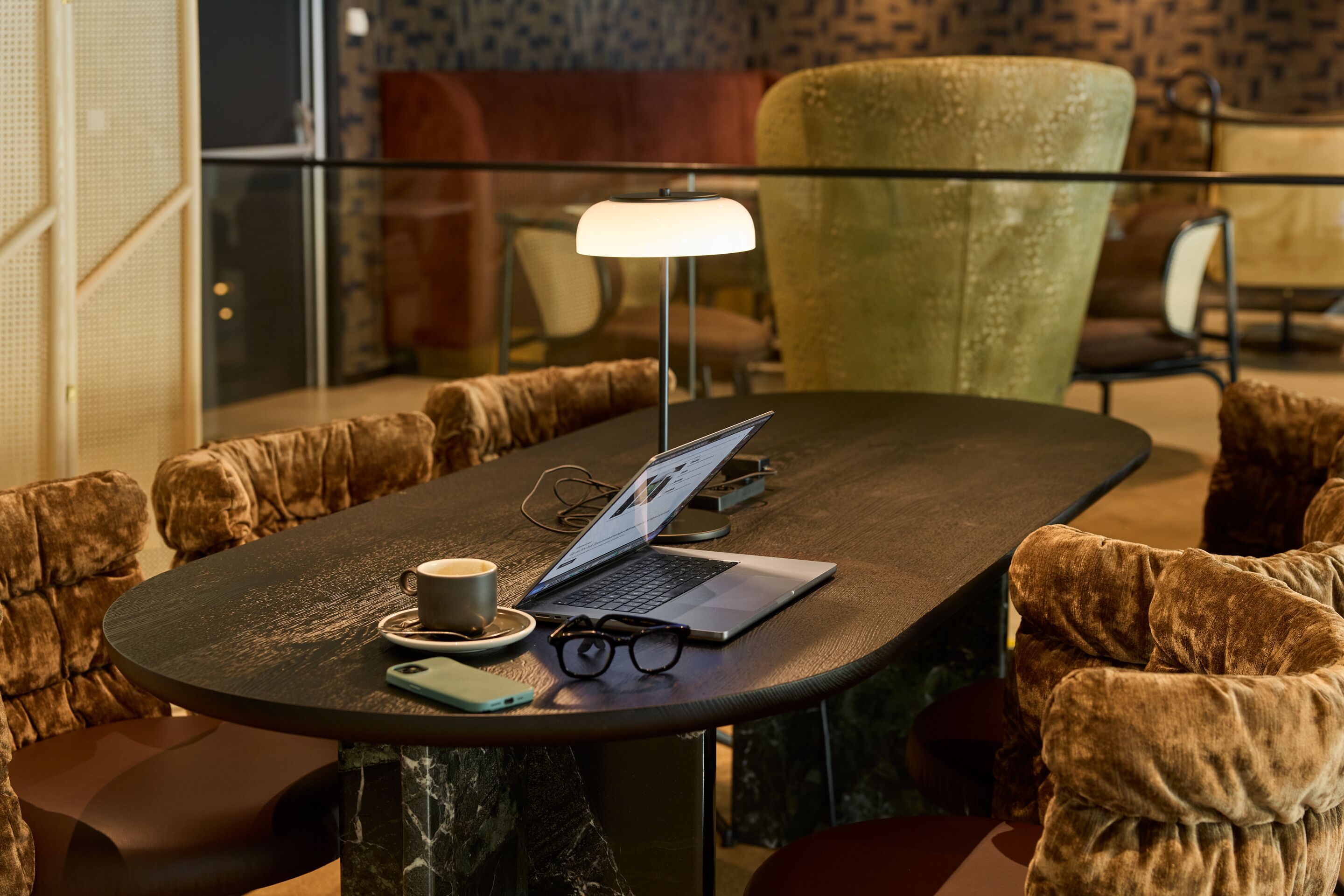 Moderner Arbeitsplatz mit Laptop, Kaffeetasse, Notizbuch und Brille auf einem dunklen Tisch, umgeben von komfortablen, gepolsterten Sesseln in einer stilvollen Hotel-Lounge.