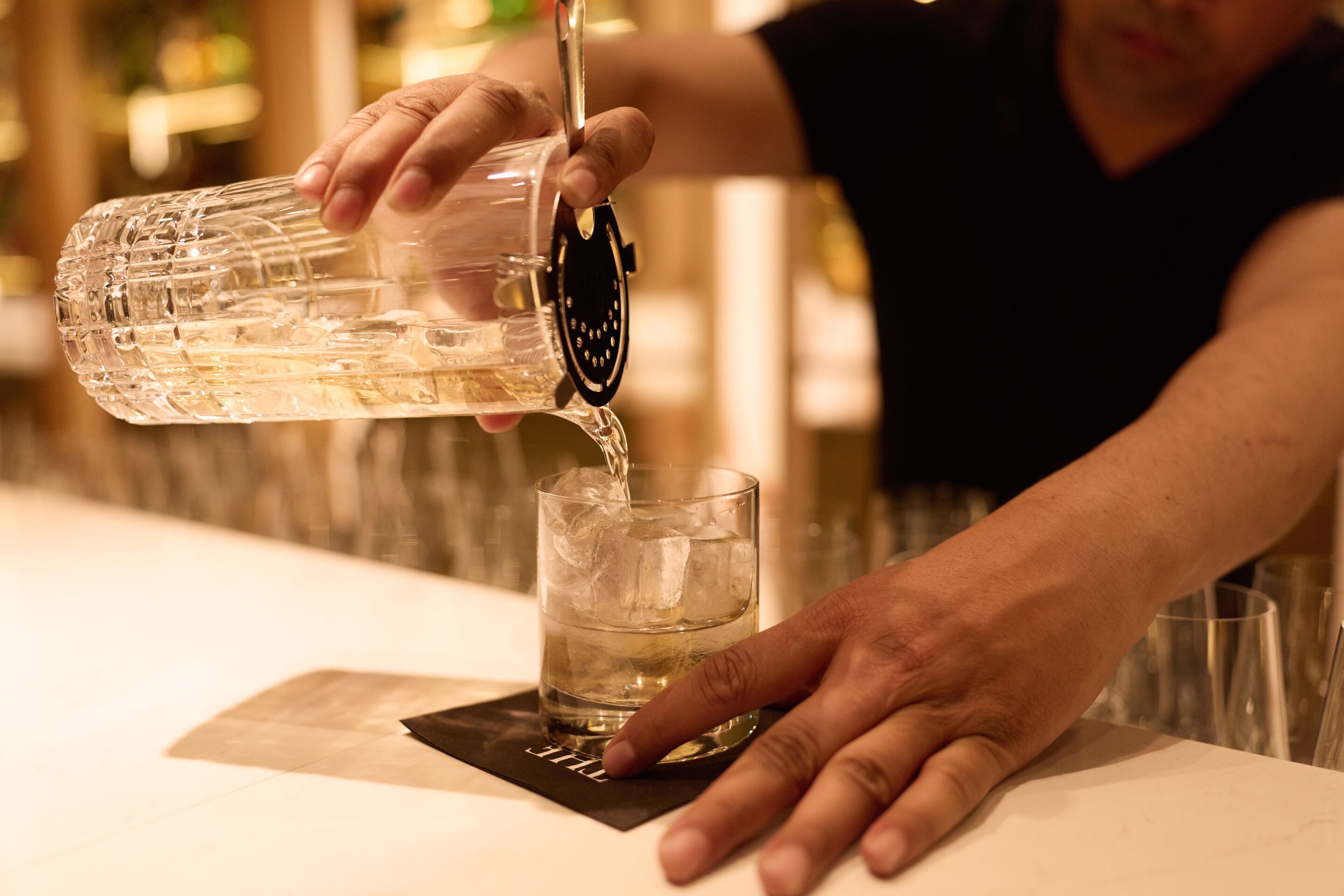 Ein Barkeeper gießt einen klaren Cocktail über Eiswürfel in einem Glas, Atmosphäre warm und einladend.