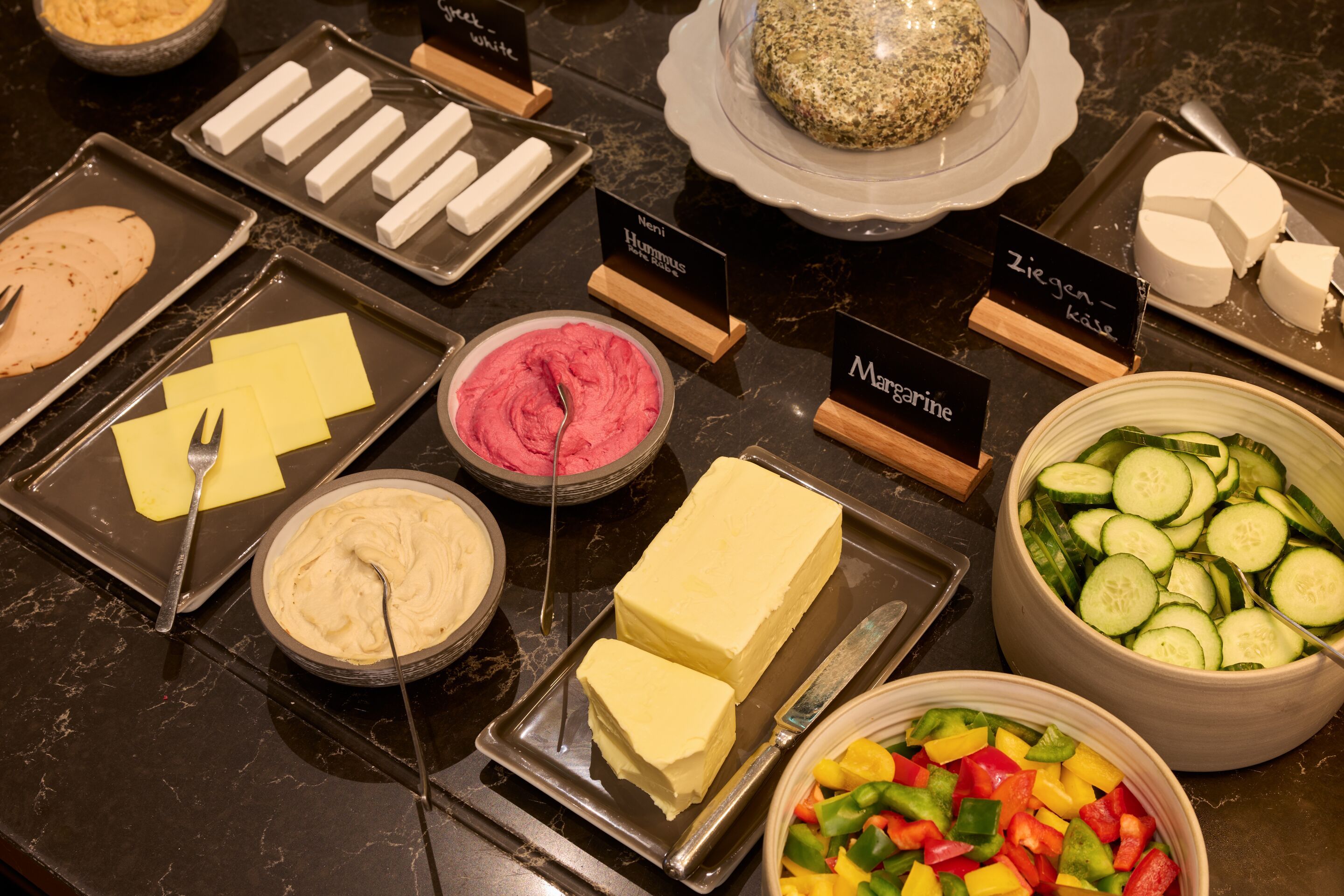 Vielfältige Aufstriche wie Margarine, Ziegenkäse und verschiedene Frischkäse, begleitet von frischem Gemüse wie Gurken- und Paprikascheiben, angerichtet auf einem Buffet für ein ausgewogenes Frühstück.