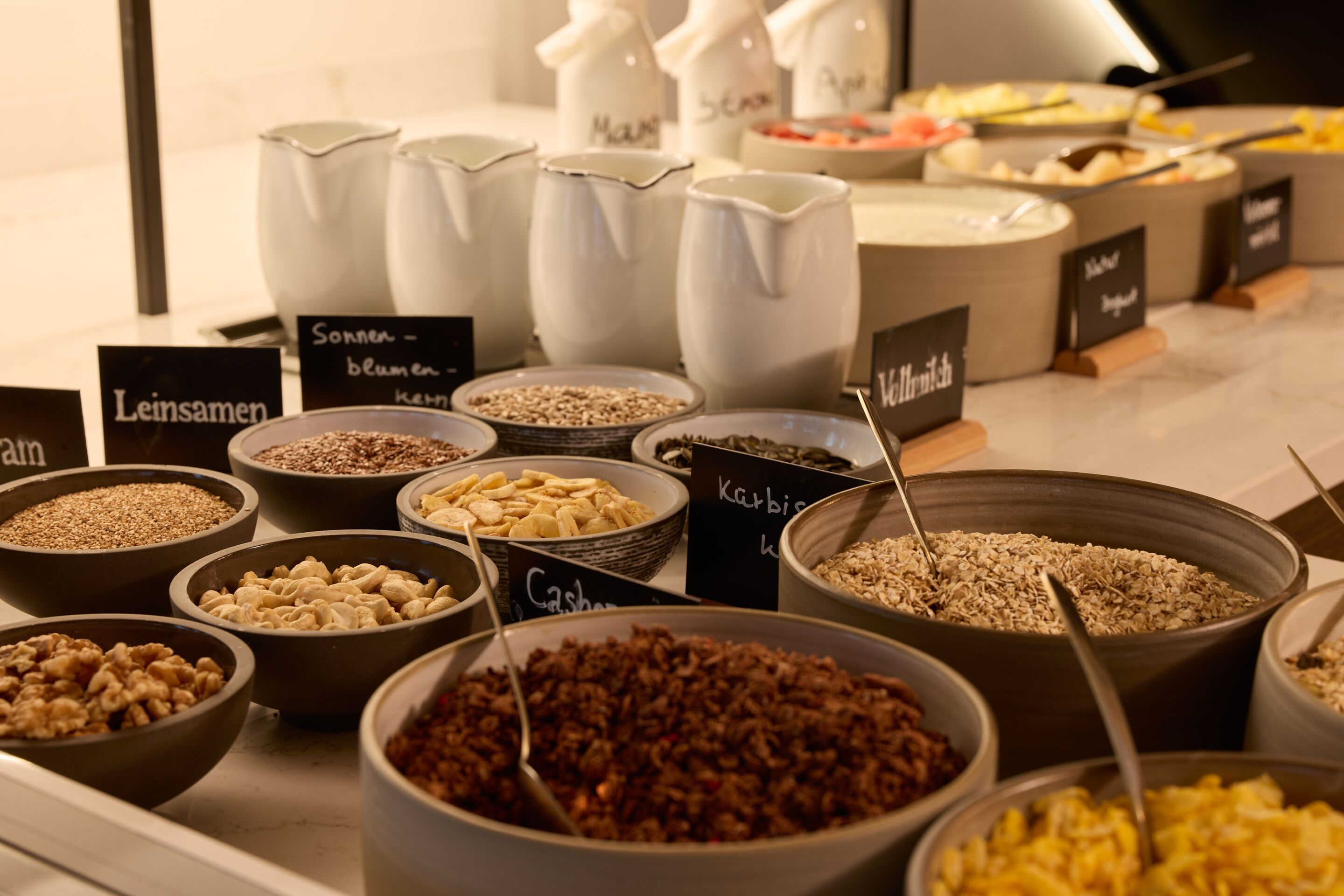 Auswahl an gesunden Frühstückszutaten wie Nüssen, Samen, Getreide und Müsli, liebevoll präsentiert in Schalen mit Beschriftungen, ideal für einen abwechslungsreichen Start in den Tag.