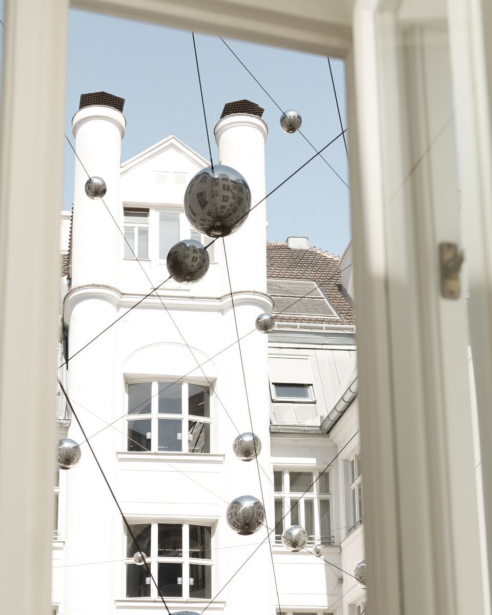 Blick aus einem Fenster auf einen Innenhof mit weißem Gebäude, in dem mehrere silberne Kugeln an dünnen Drähten als moderne Kunstinstallation hängen.