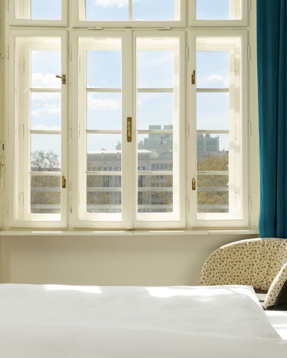 Helles Hotelzimmer mit großem Fenster, das Tageslicht hereinlässt. Sicht auf Stadtgebäude und klare Himmelskulisse. Gemütlicher Sessel neben dem Fenster, weiße Bettdecke im Vordergrund. Ruhe und helle, freundliche Atmosphäre.