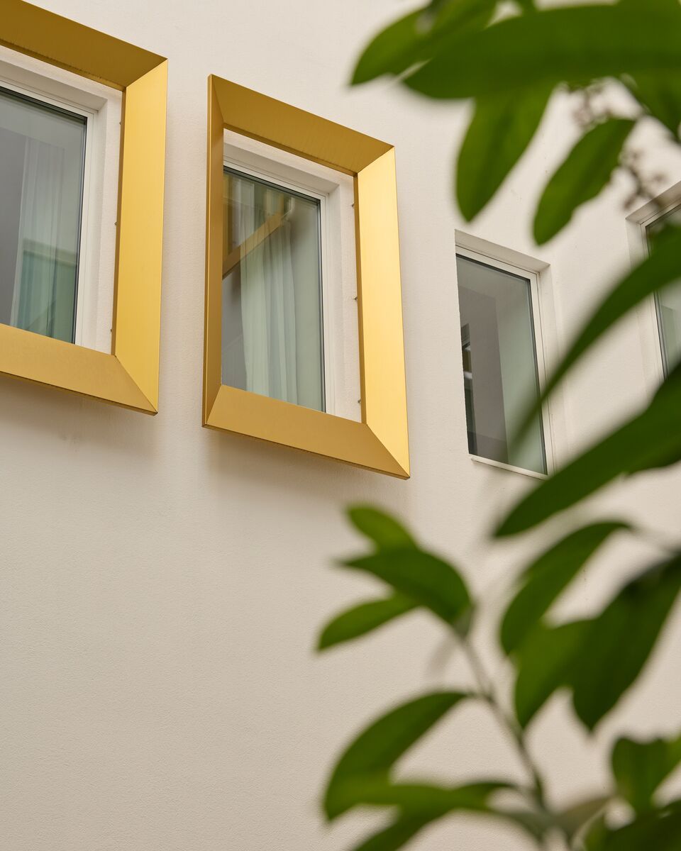 Drei moderne Fenster mit goldfarbenen Rahmen an einer weißen Fassade, teilweise von grünen Pflanzen im Vordergrund umgeben, wirken elegant und einladend.