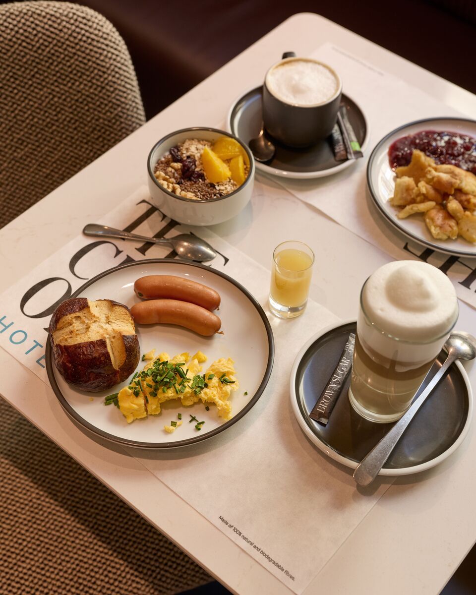 Frühstückstisch mit Rührei, zwei Würstchen, Brezel, Müsli mit Früchten, Apfelmus mit Zimt, Espresso, Latte Macchiato und einem Glas Orangensaft. Gemütliche Atmosphäre für einen energiereichen Start in den Tag.