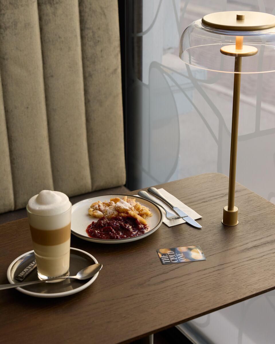 Kaffeetisch mit Latte Macchiato und Teller mit Kirschenpfannkuchen, neben modernem goldfarbenen Tischlampenlicht und bequemer Polsterbank, gemütliche Atmosphäre für entspannte Momente im Hotel.