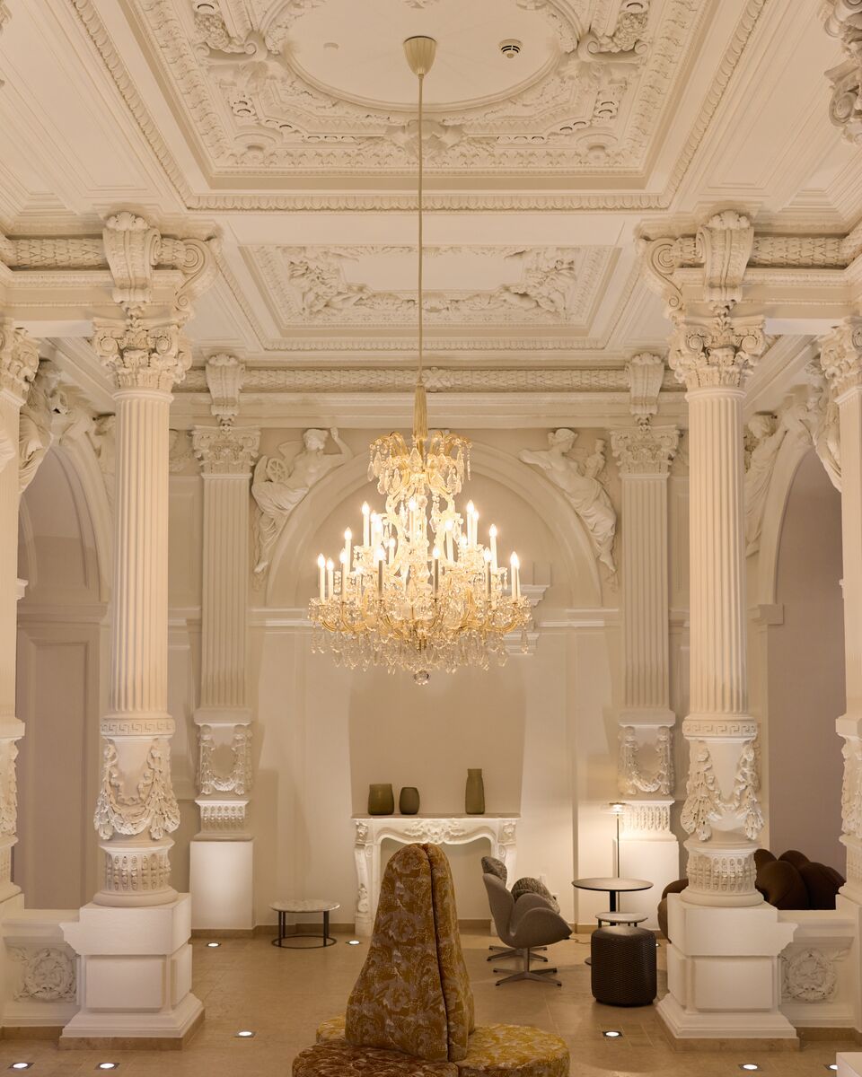Prächtiger Raum mit kunstvoll verzierten Säulen und Stuckdecken, dominiert von einem eleganten, goldenen Kronleuchter, der warmes Licht spendet. Gemütliche Sitzgelegenheiten und dezente Beleuchtung schaffen eine einladende, luxuriöse Atmosphäre.