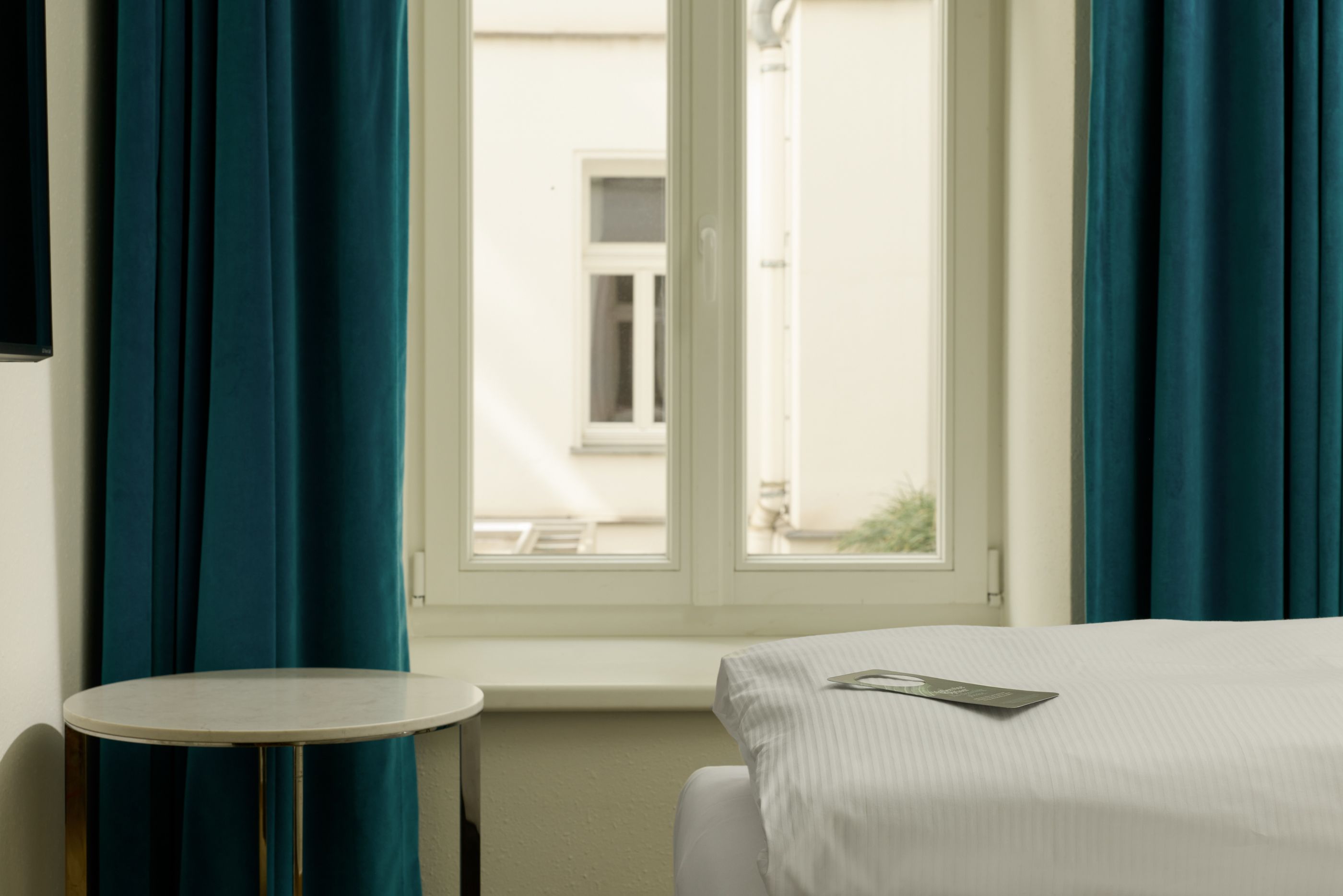 Hotel The Cloud One Wien-Staatsoper Hell eingerichtetes Hotelzimmer mit großem Fenster, weißen Wänden und dicken blauen Vorhängen. Neben dem Fenster ein kleines rundes Tischchen, davor ein gemütlich gemachtes Bett mit weißer Bettwäsche.