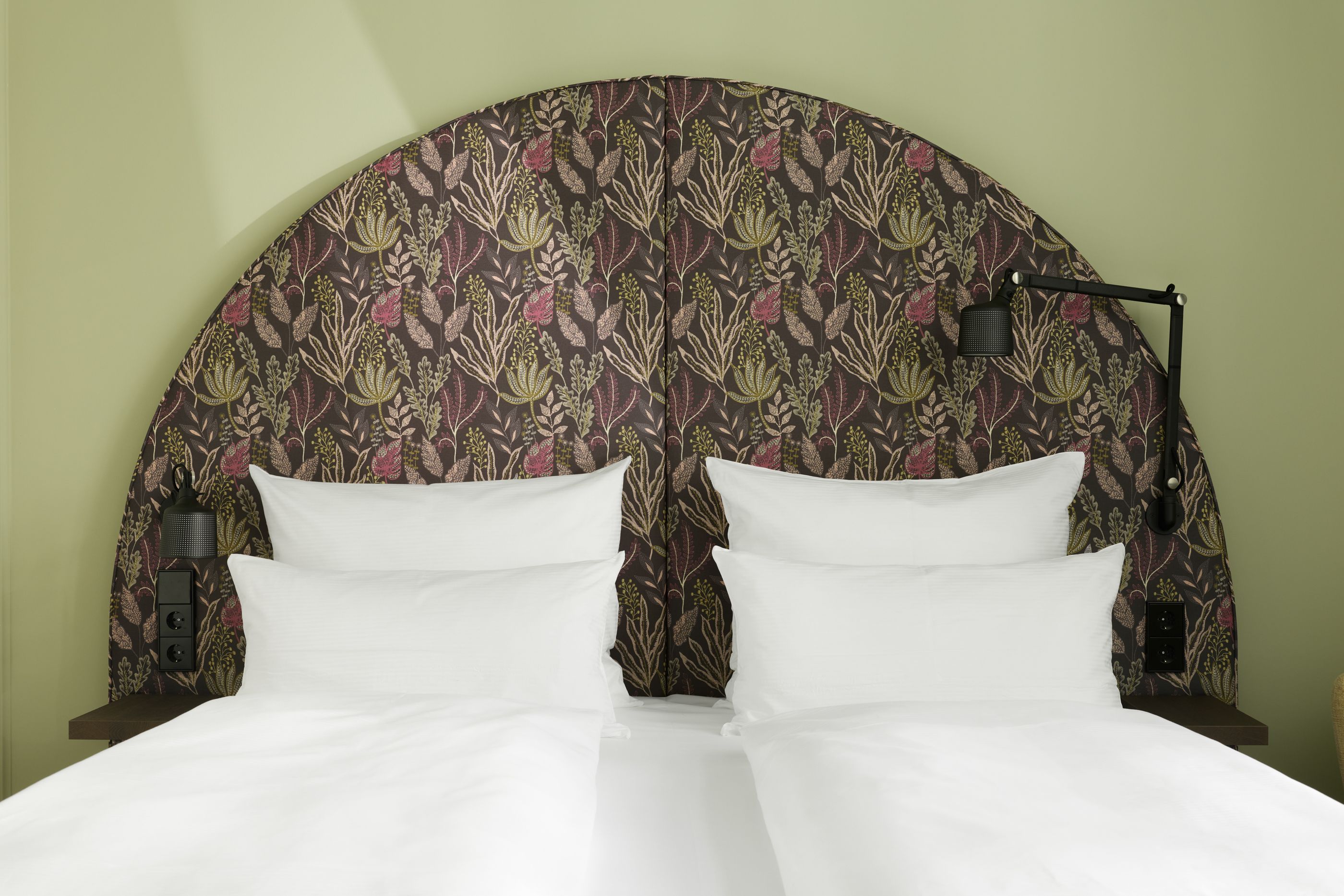 Hotel The Cloud One Wien-Staatsoper Doppelbett mit weißer Bettwäsche vor einem großen, abgerundeten Kopfteil mit floralem Muster, umgeben von sanfter Beleuchtung.