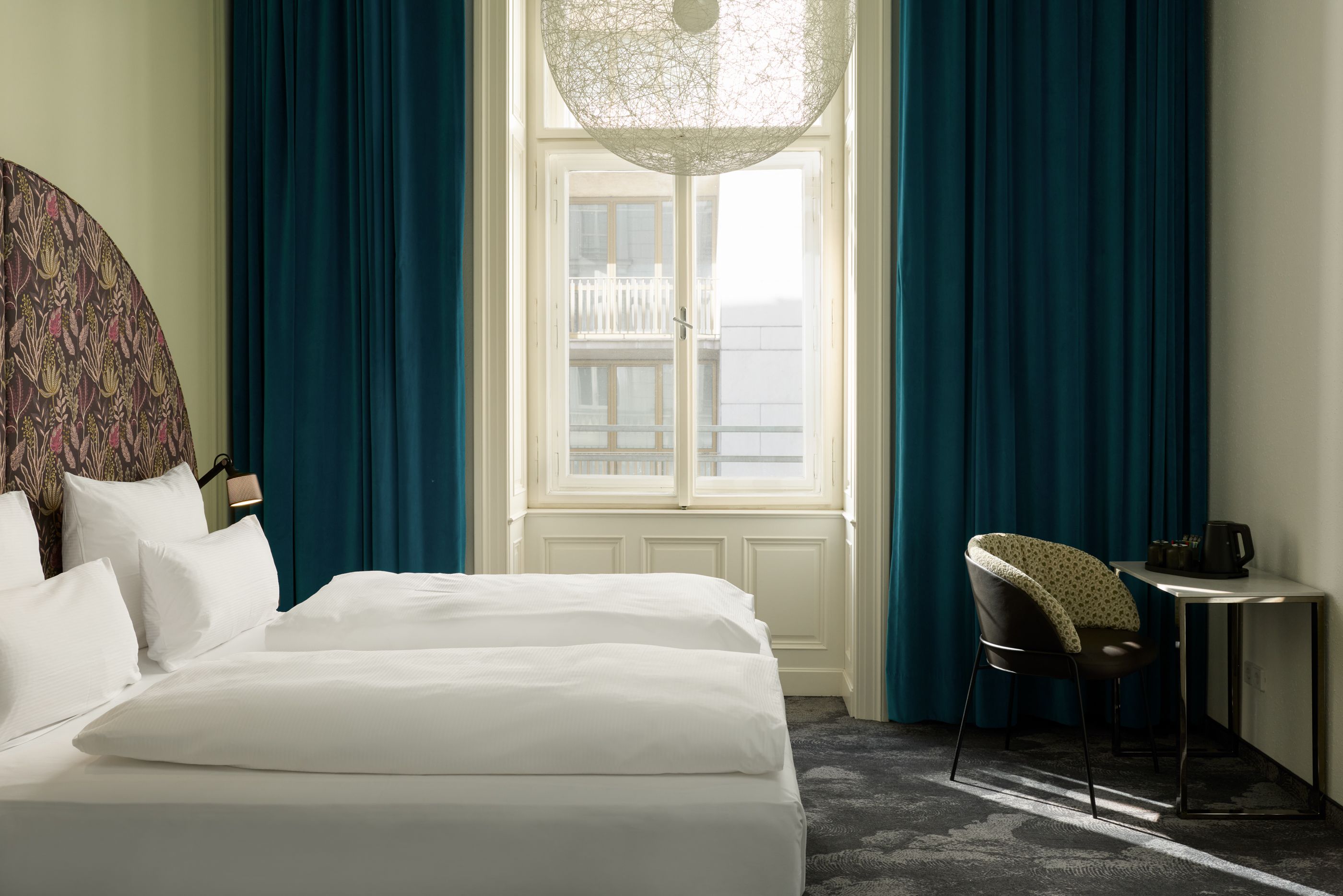 Hotel The Cloud One Wien-Staatsoper Helles Hotelzimmer mit zwei modernen Einzelbetten, großem Fenster mit Tageslicht, blauem Vorhang und stilvollem Arbeitsplatz mit Stuhl. Atmosphäre wirkt ruhig und einladend.