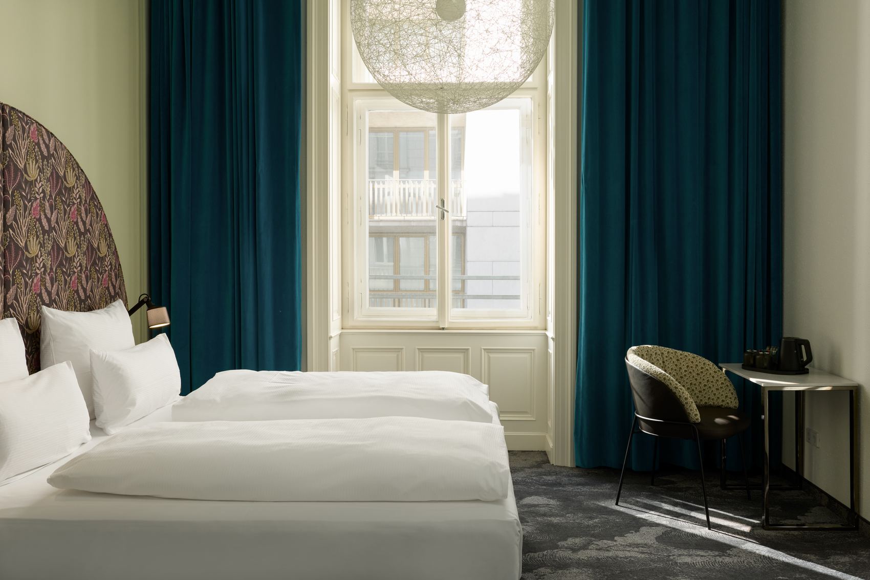 Hotel The Cloud One Wien-Staatsoper Helles Hotelzimmer mit zwei modernen Einzelbetten, großem Fenster mit Tageslicht, blauem Vorhang und stilvollem Arbeitsplatz mit Stuhl. Atmosphäre wirkt ruhig und einladend.