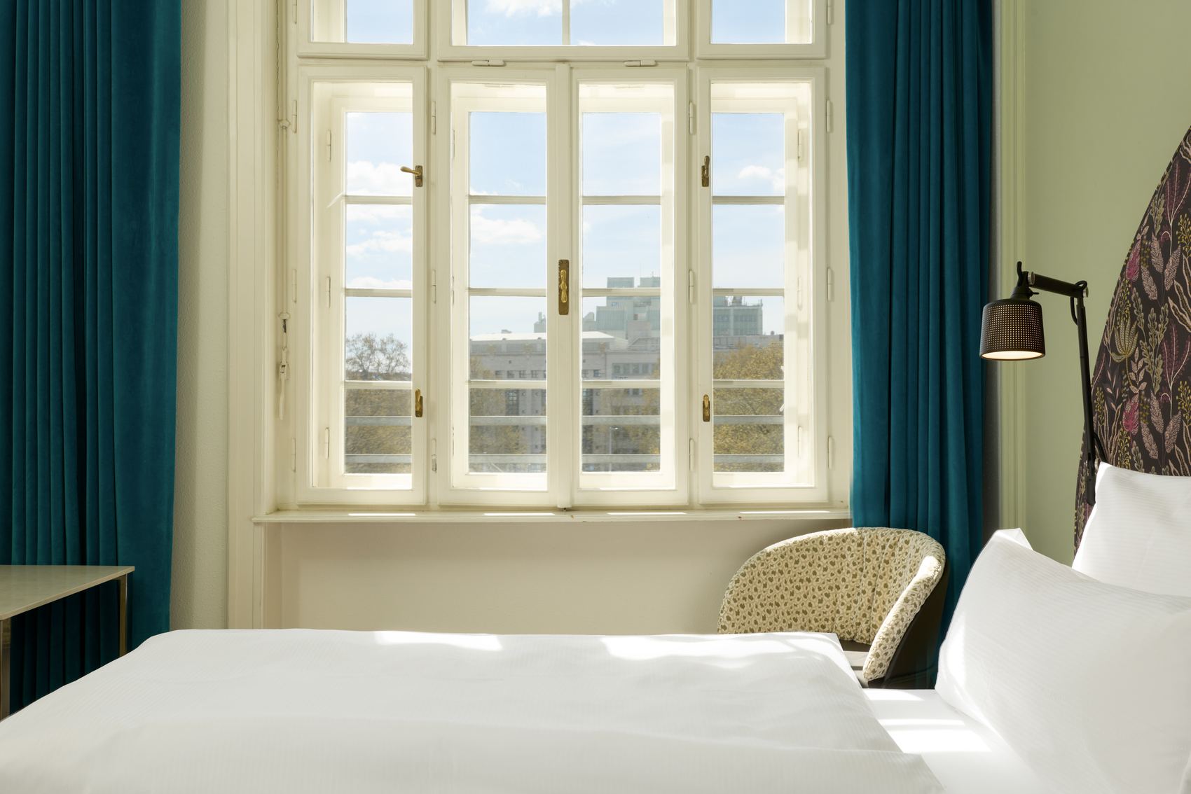 Hotel The Cloud One Wien-Staatsoper Helles Hotelzimmer mit großem Fenster, das Tageslicht hereinlässt. Weiß bezogenes Bett, gemütlicher Sessel und blaue Vorhänge schaffen eine ruhige und einladende Atmosphäre. Blick auf eine Stadtlandschaft durch das Fenster.