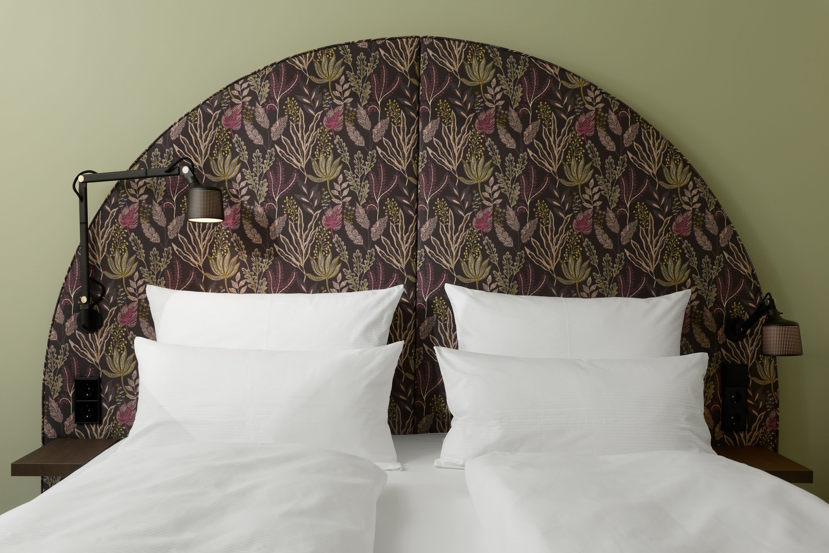 Hotel The Cloud One Wien-Staatsoper Ein gemütliches Doppelbett mit weißen Bettlaken und Kissen, davor ein großer, halbkreisförmiger, gemusterter Kopfteil in dunklen Naturtönen, seitlich zwei verstellbare Leselampen an der Wand.