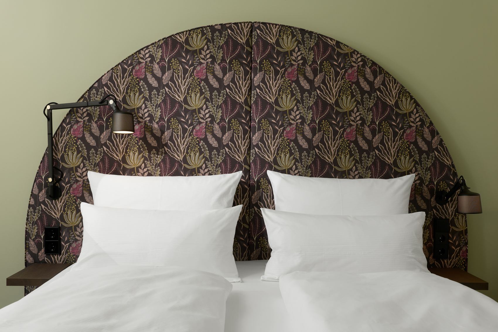 Hotel The Cloud One Wien-Staatsoper Ein gemütliches Doppelbett mit weißen Bettlaken und Kissen, davor ein großer, halbkreisförmiger, gemusterter Kopfteil in dunklen Naturtönen, seitlich zwei verstellbare Leselampen an der Wand.