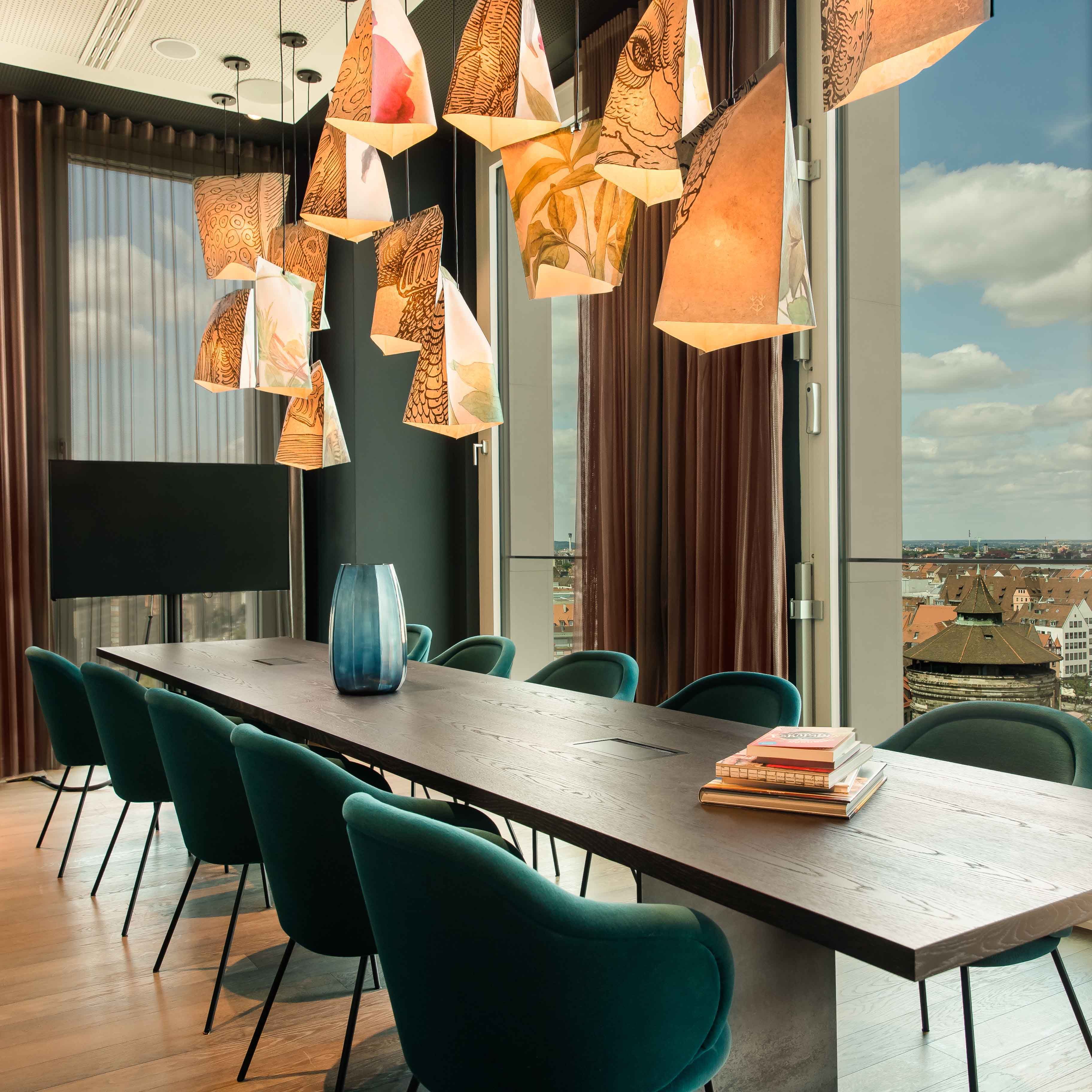 Motel One Konferenzraum mit langem Holztisch, dunkelgrünen Stühlen und auffälligen kunstvollen Hängelampen. Große Fenster bieten Tageslicht und Blick auf Stadt und Himmel. 