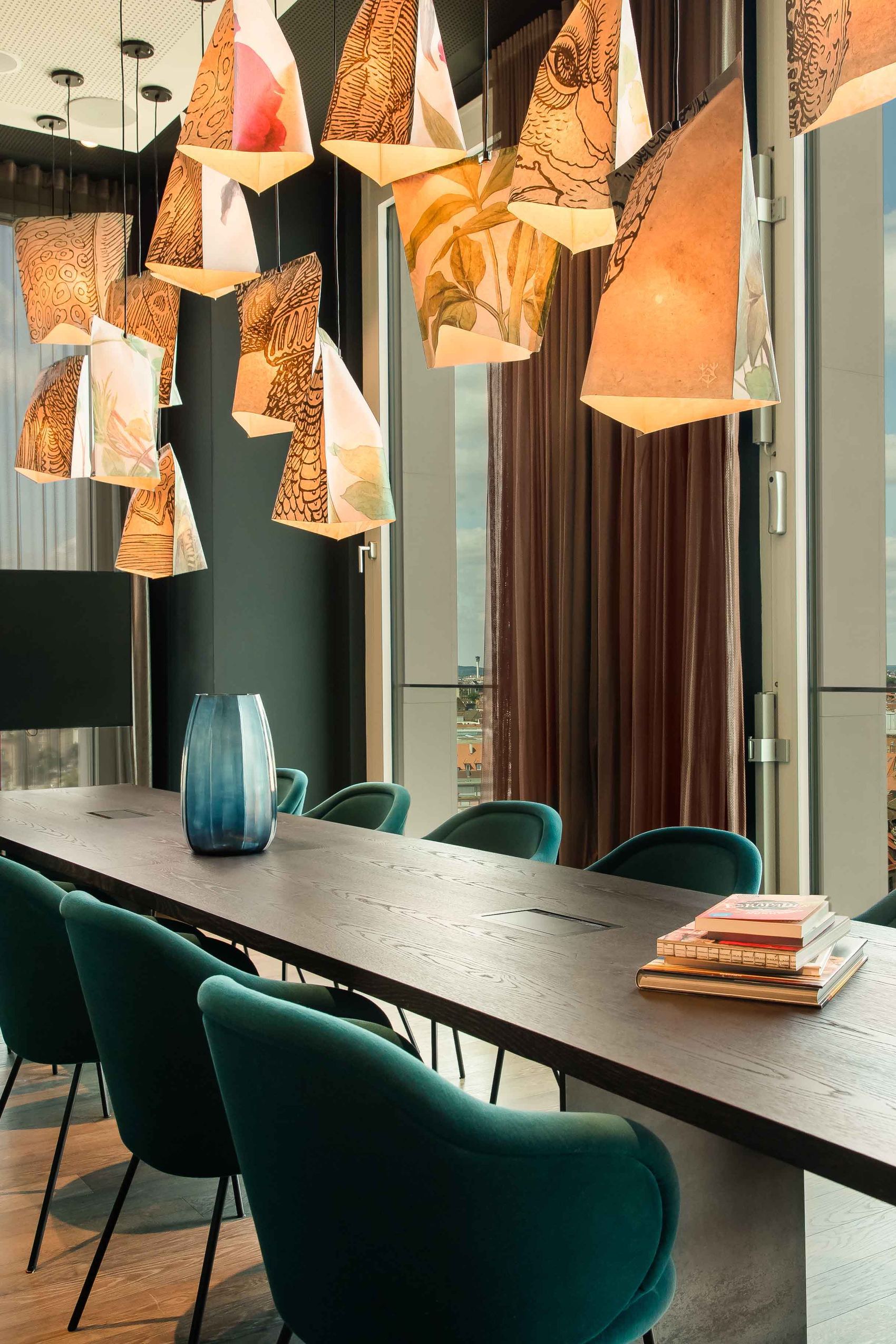 Motel One Konferenzraum mit langem Holztisch, dunkelgrünen Stühlen und auffälligen kunstvollen Hängelampen. Große Fenster bieten Tageslicht und Blick auf Stadt und Himmel.