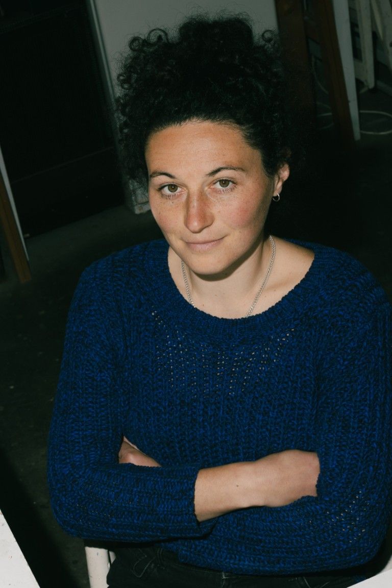 Porträt einer Frau mit lockigem, dunklem Haar, die lächelnd und mit verschränkten Armen in einem gemütlichen blauen Pullover sitzt.