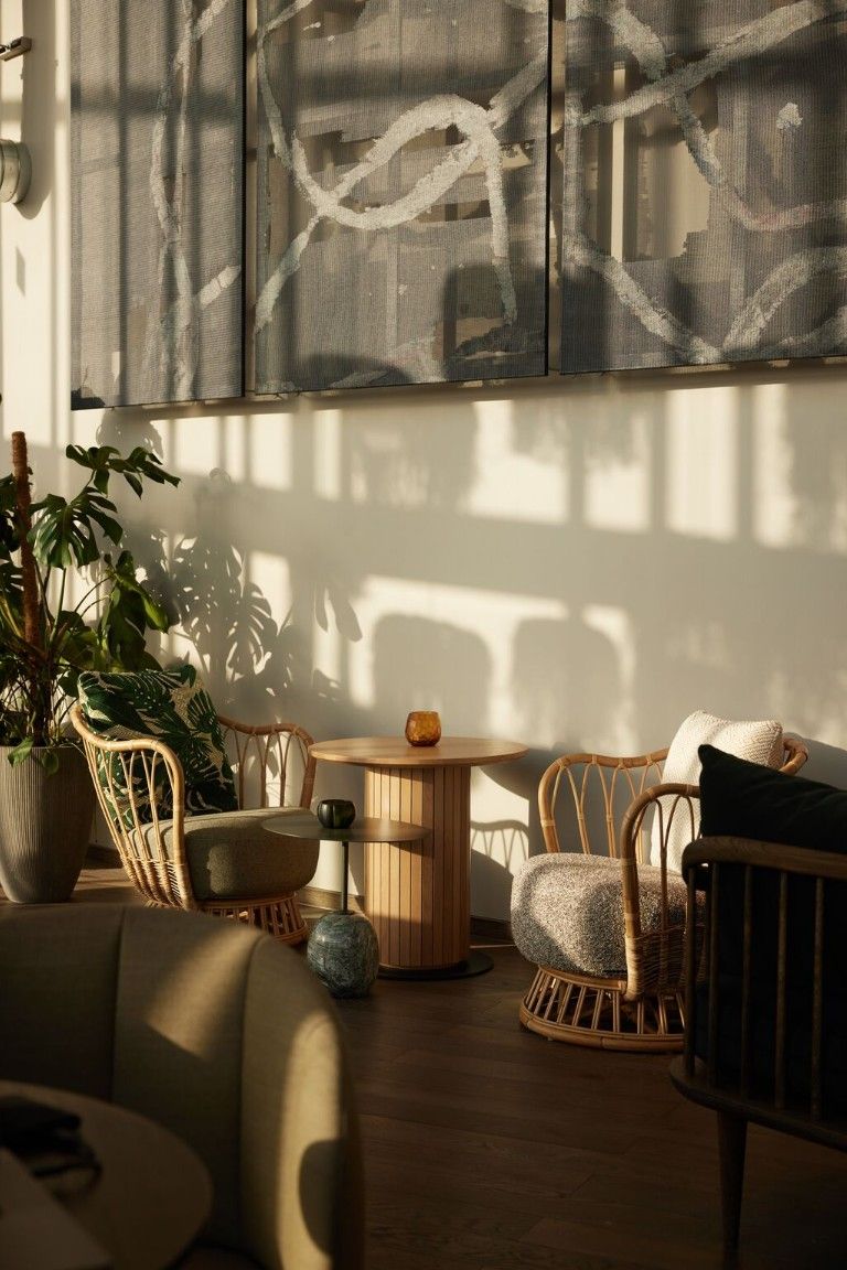 Helles, gemütliches Lounge-Eck mit Rattanstühlen, einem Holzbeistelltisch und Pflanzen. Sanftes Sonnenlicht und Schatten schaffen eine entspannte, warme Atmosphäre.