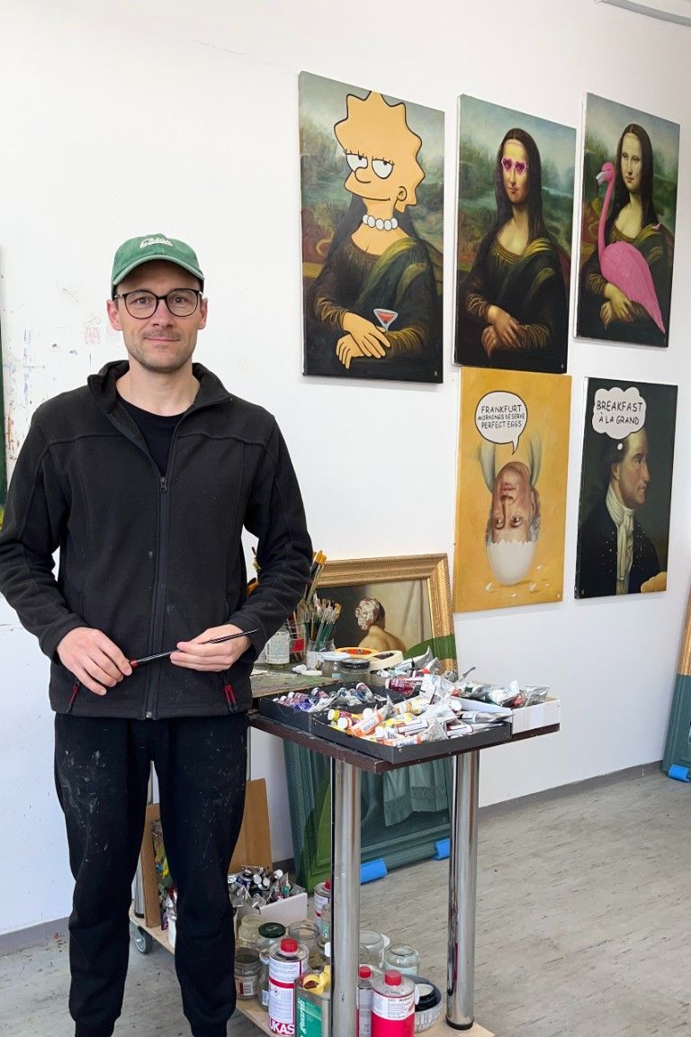 Künstler steht lächelnd in seinem Atelier vor einer Wand mit humorvollen Mona-Lisa-Variationen, darunter eine mit Lisa Simpson und eine mit einem Flamingo. Vor ihm Tisch mit Malutensilien und Farben.