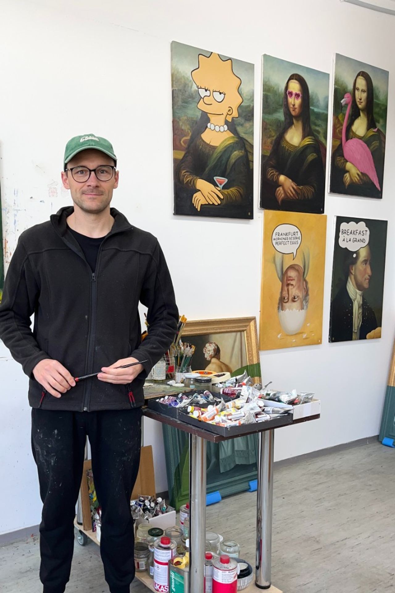 Künstler steht lächelnd in seinem Atelier vor einer Wand mit humorvollen Mona-Lisa-Variationen, darunter eine mit Lisa Simpson und eine mit einem Flamingo. Vor ihm Tisch mit Malutensilien und Farben.