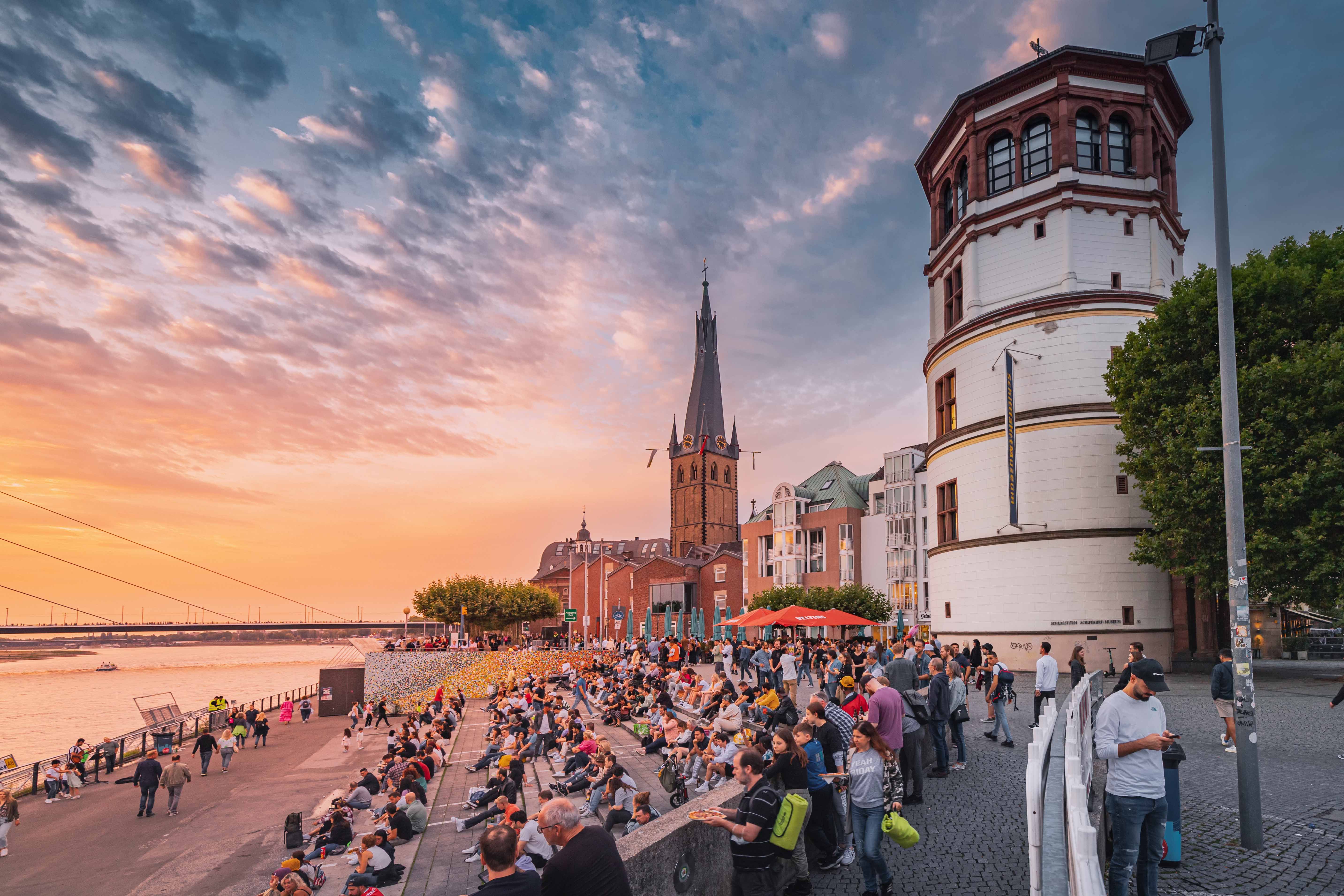 Menschen sitzen an einem Flussufer bei Sonnenuntergang, mit Blick auf historische Gebäude und einen Wasserturm, die eine entspannte, lebendige Atmosphäre am Wasser schaffen.