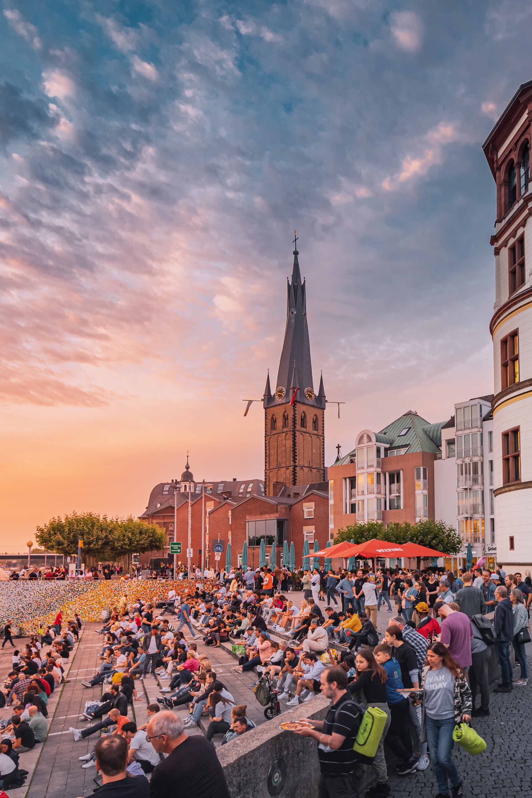Menschen sitzen an einem Flussufer bei Sonnenuntergang, mit Blick auf historische Gebäude und einen Wasserturm, die eine entspannte, lebendige Atmosphäre am Wasser schaffen.