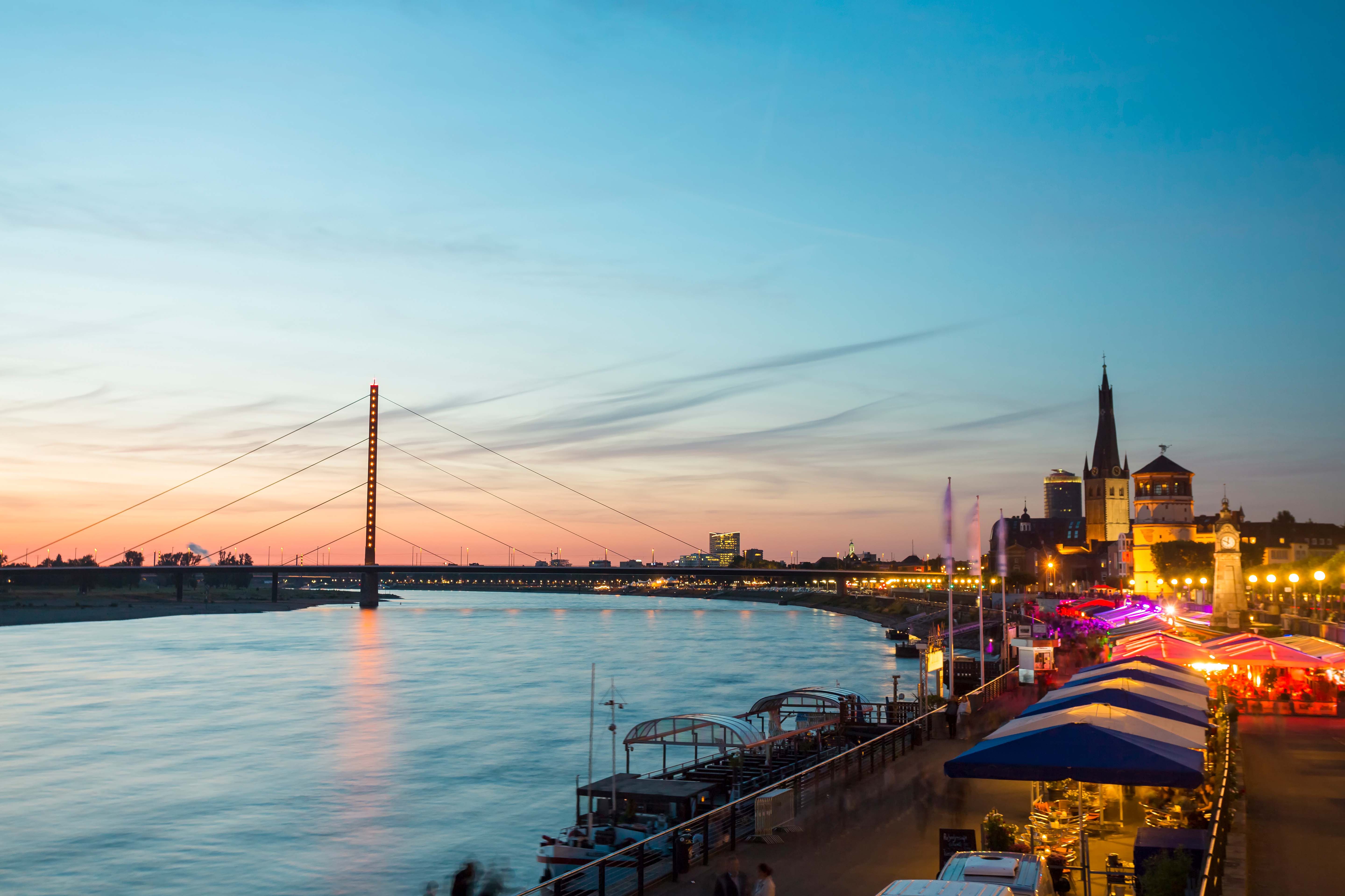Blick auf die beleuchtete Rheinuferpromenade bei Dämmerung mit Blick auf die Schwebefähre und historische Kirchen im Hintergrund, atmosphärisch und einladend für Abendspaziergänge und kulturelle Erlebnisse.