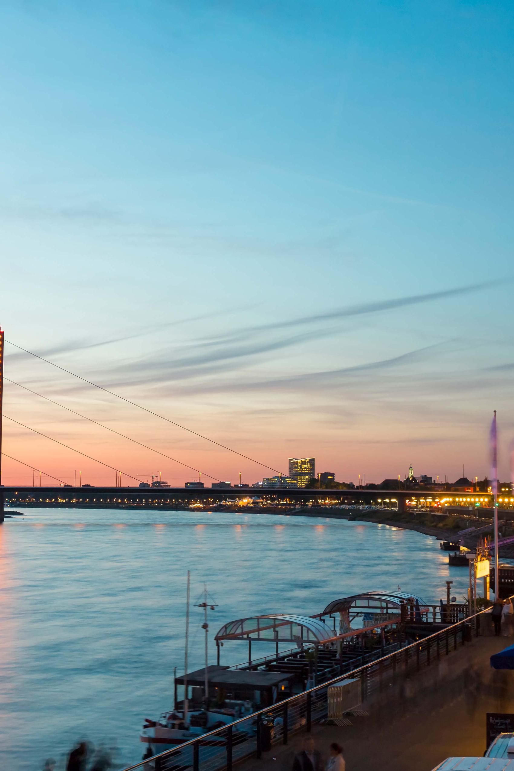 Blick auf die beleuchtete Rheinuferpromenade bei Dämmerung mit Blick auf die Schwebefähre und historische Kirchen im Hintergrund, atmosphärisch und einladend für Abendspaziergänge und kulturelle Erlebnisse.