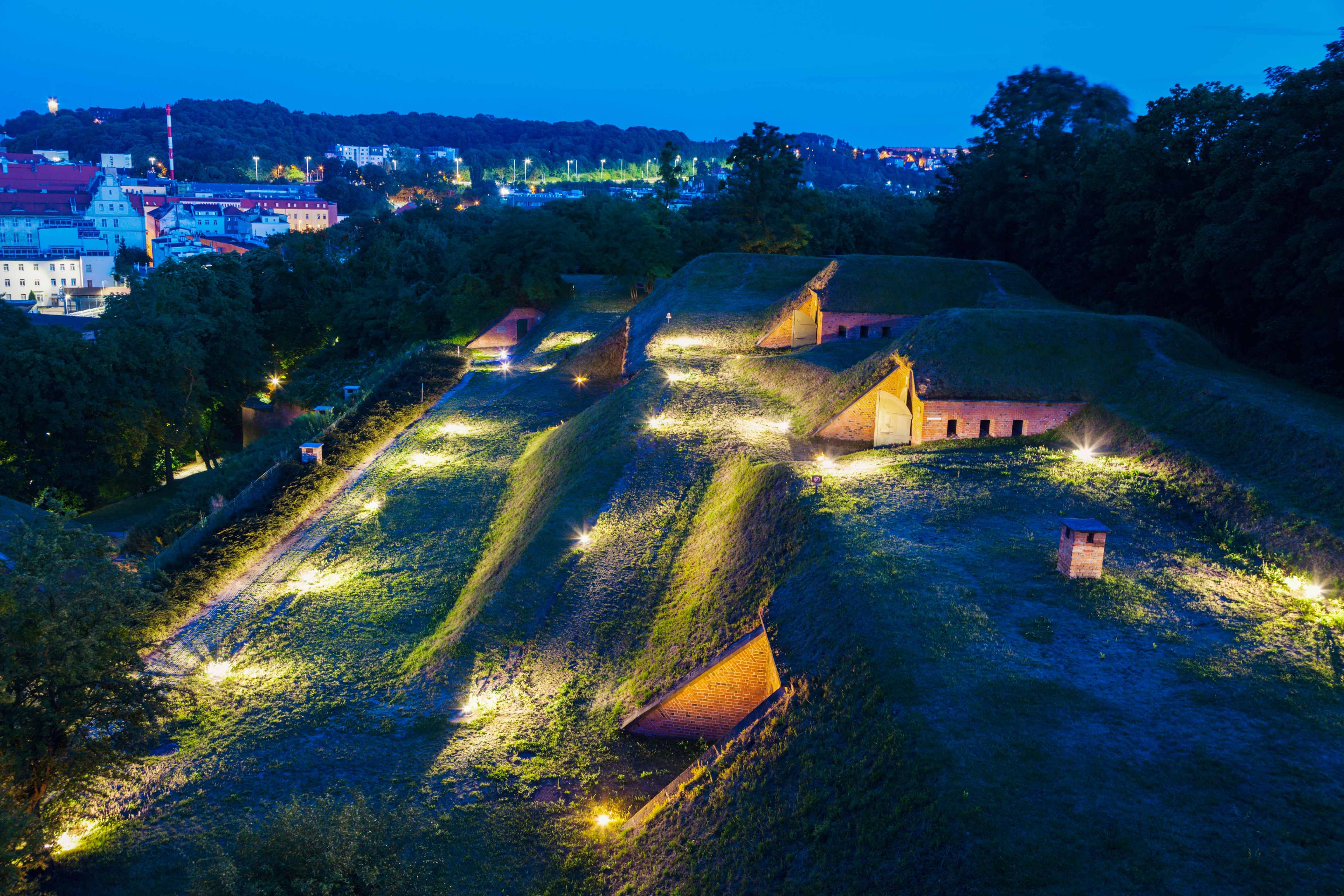 Beleuchtete Hügelburg mit Grasdächern bei Nacht, umgeben von Bäumen, im Hintergrund eine erleuchtete Stadtlandschaft.