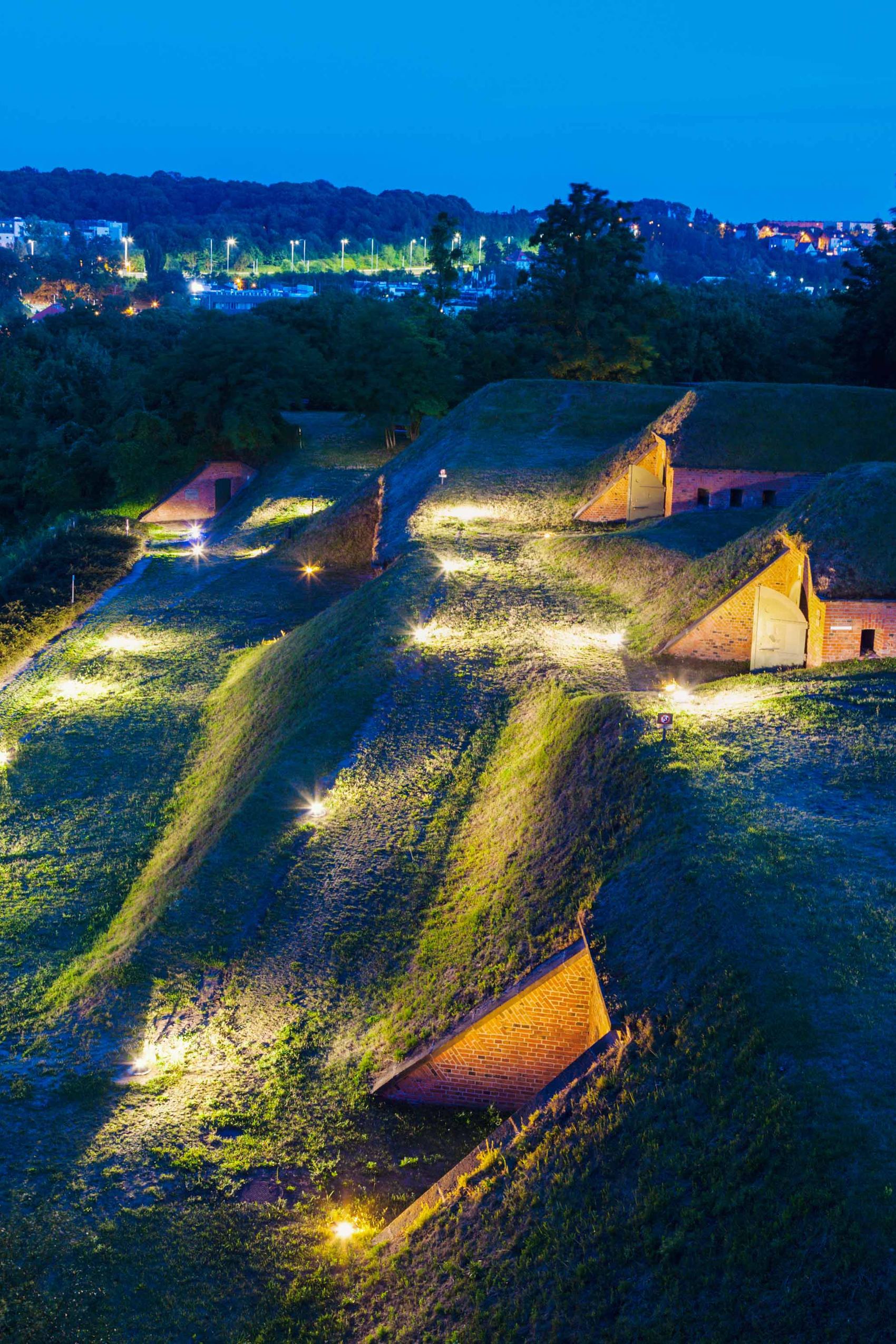 Beleuchtete Hügelburg mit Grasdächern bei Nacht, umgeben von Bäumen, im Hintergrund eine erleuchtete Stadtlandschaft.