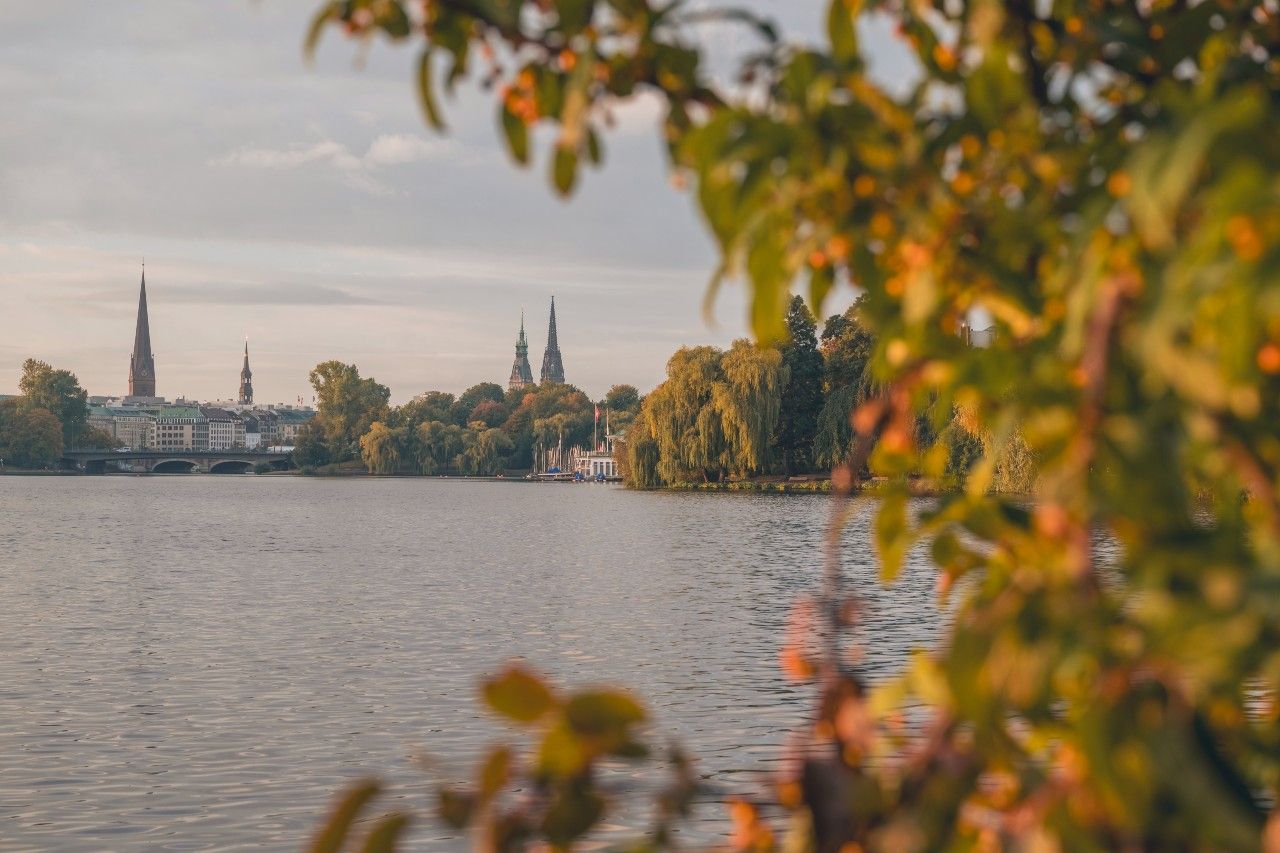 Blick auf einen ruhigen See mit grüner Ufervegetation, im Hintergrund sind mehrere markante Kirchtürme einer Stadt zu sehen, unter warmem, weichem Licht eines späten Nachmittags.