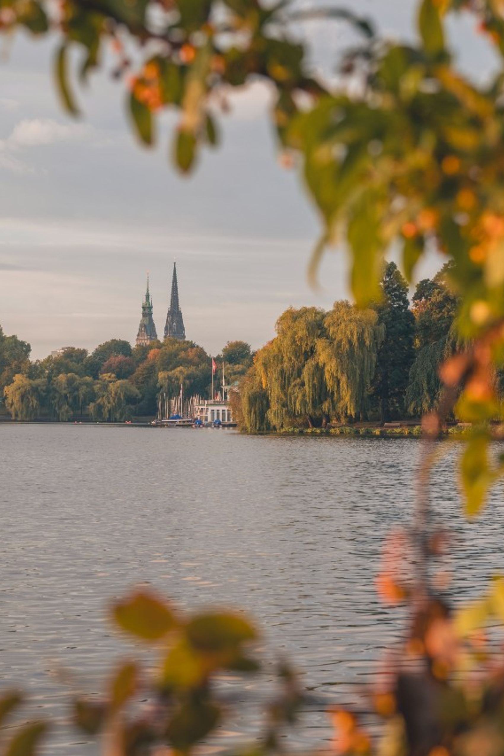Blick auf einen ruhigen See mit grüner Ufervegetation, im Hintergrund sind mehrere markante Kirchtürme einer Stadt zu sehen, unter warmem, weichem Licht eines späten Nachmittags.