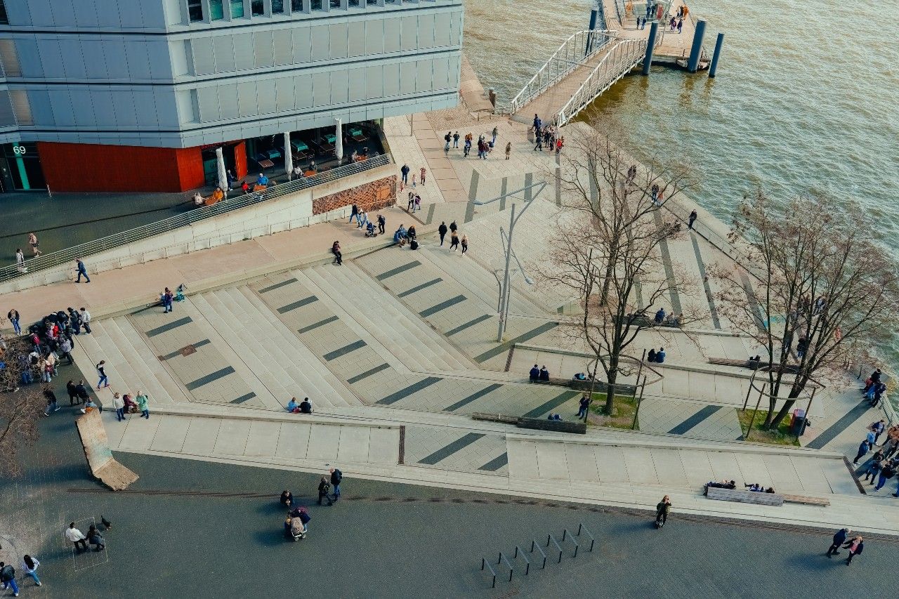 Blick auf einen modernen, terrassenförmigen Platz an einem Flussufer mit Bäumen, Spaziergängern und einer Anlegestelle, ideal für entspannte Pausen und Ausblicke aufs Wasser.