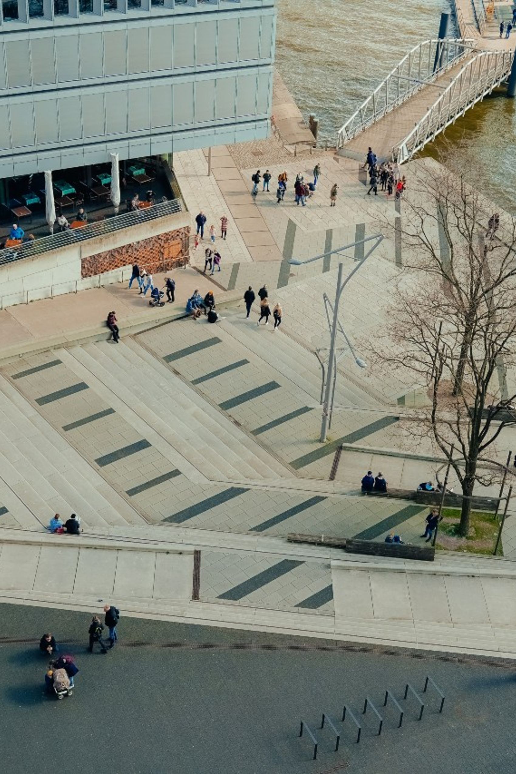 Blick auf einen modernen, terrassenförmigen Platz an einem Flussufer mit Bäumen, Spaziergängern und einer Anlegestelle, ideal für entspannte Pausen und Ausblicke aufs Wasser.