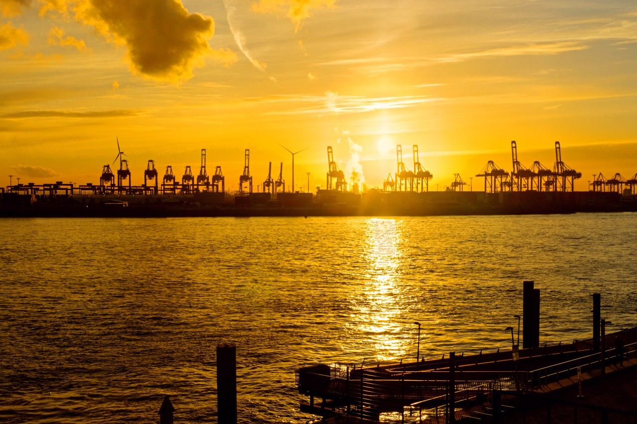 Sonnenuntergang über einem Industriehafen mit Kränen und Windrädern, die am Ufer stehen, reflektiert goldenes Licht auf dem ruhigen Wasser.